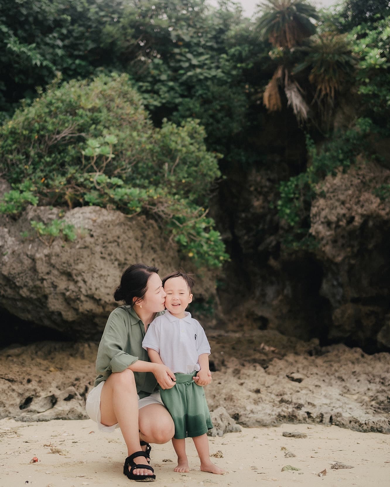 岩と緑に囲まれた沖縄の浜辺で、幼い子どもの頬にキスをする母親