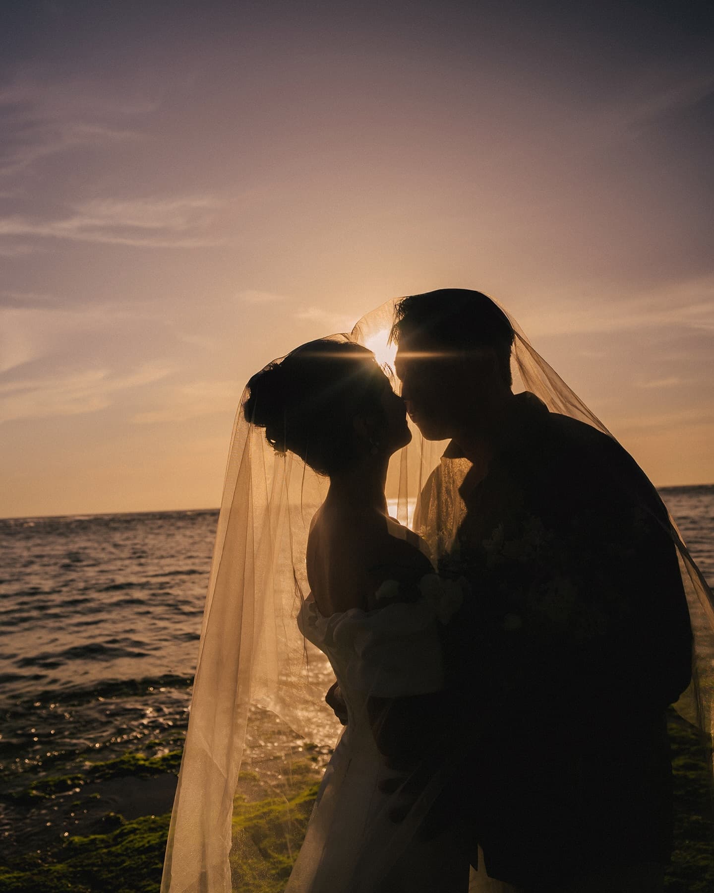 Silhouetted couple kissing under a flowing veil at sunset on an Okinawa beach