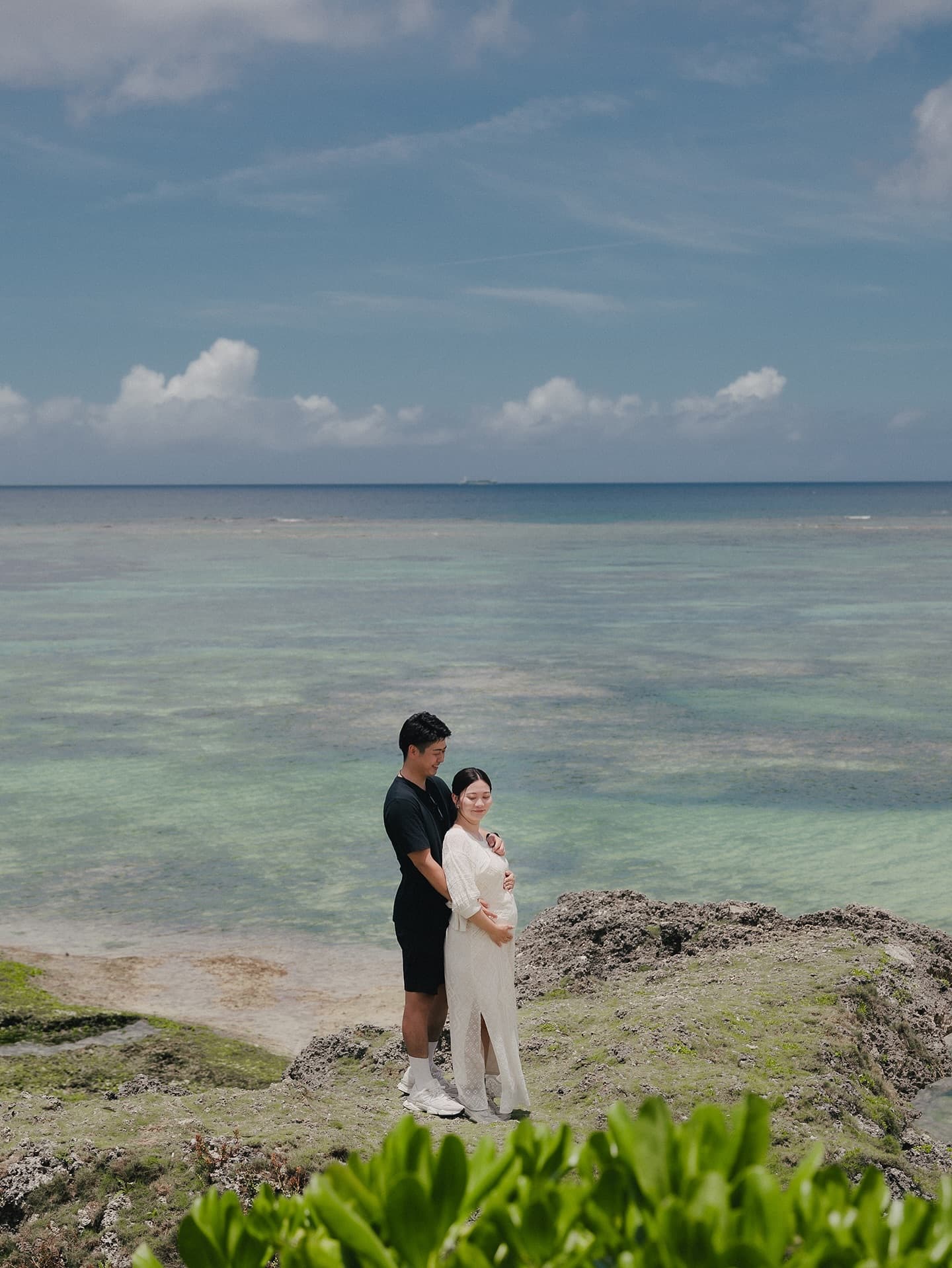 エメラルドの浅瀬を望む岩場でお腹に手を添え寄り添う妊婦と夫、沖縄