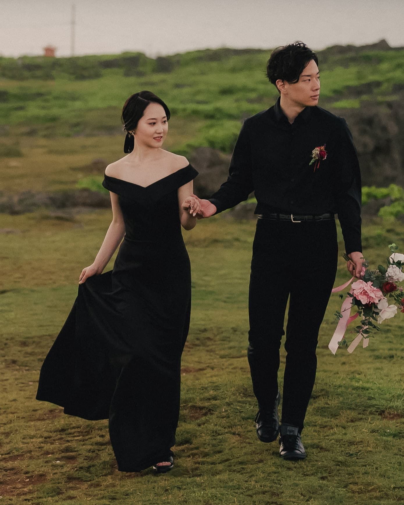 Couple walking hand in hand across a grassy field, bride in an off-shoulder black gown