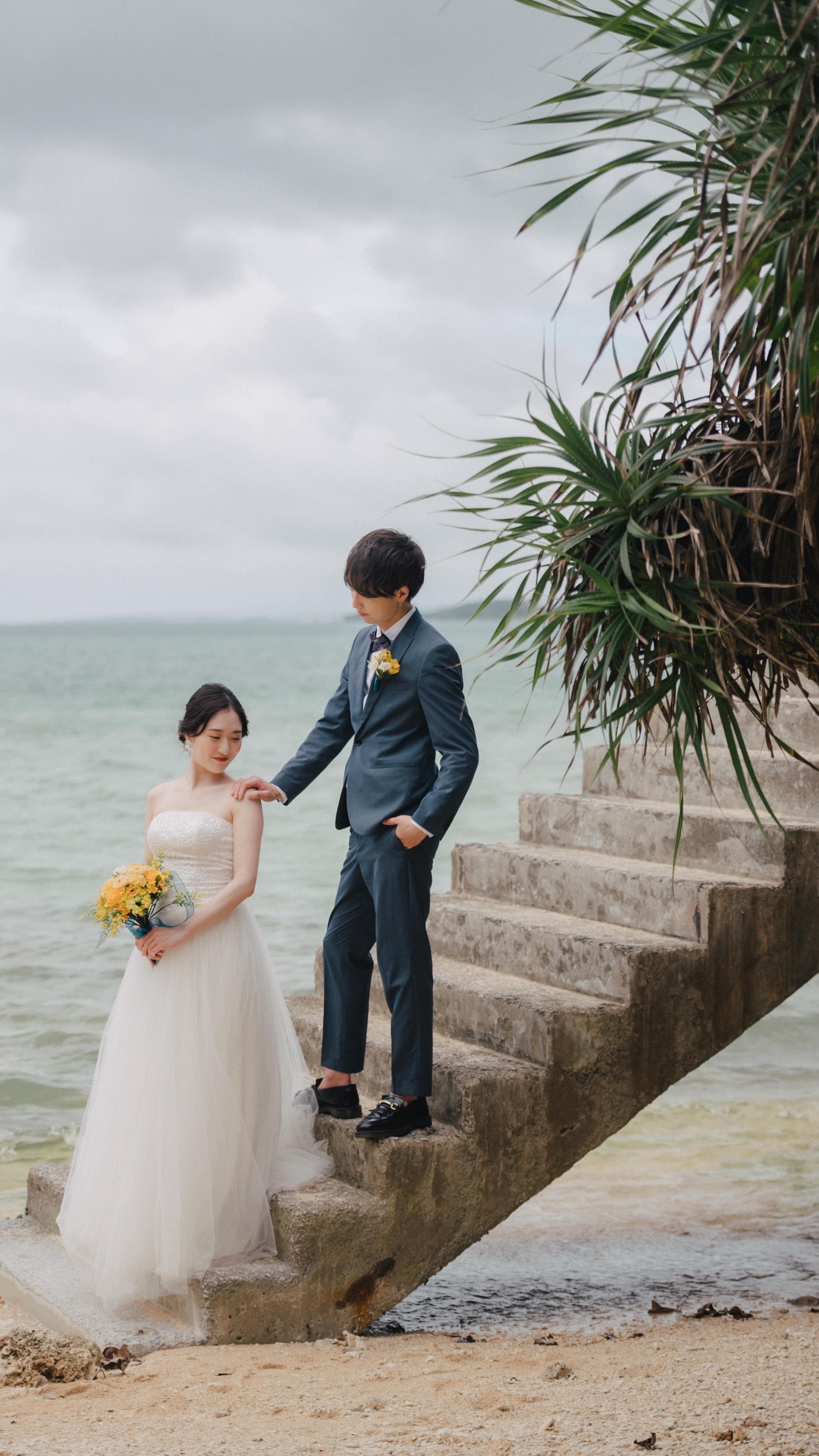 曇り空の海辺の石段で、黄色いブーケの花嫁に寄り添う新郎。沖縄。