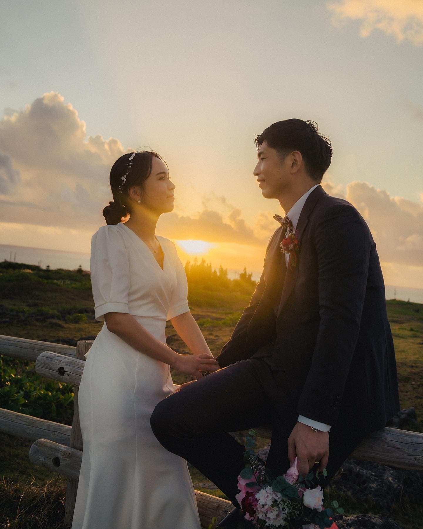 夕焼け空の下、木の柵に腰掛けて見つめ合う沖縄の新郎新婦
