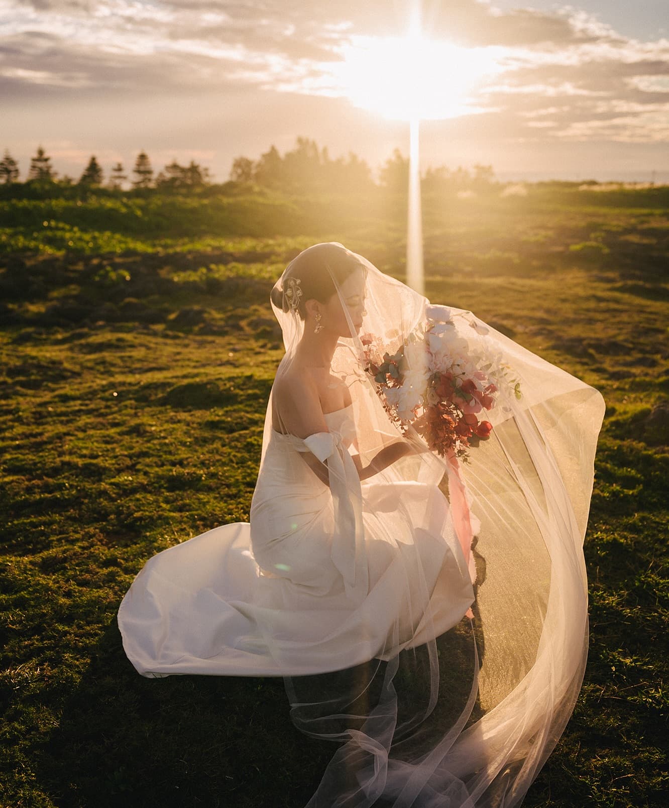 Bride with a long veil and bouquet seated on a grassy field at sunset in Okinawa