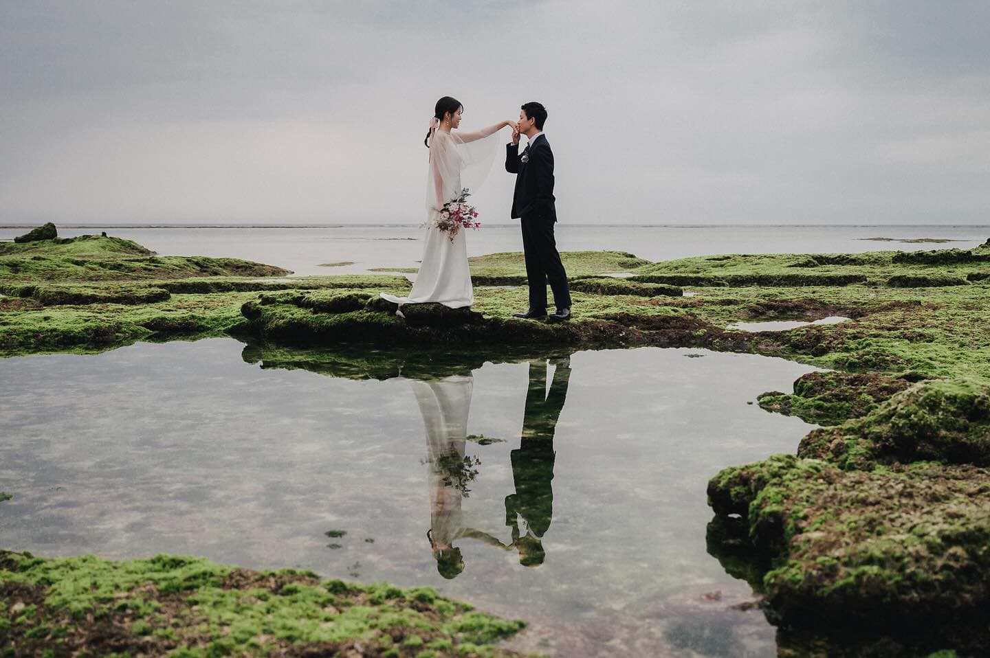 Couple mirrored in a tide pool among mossy rocks under an overcast Okinawa sky