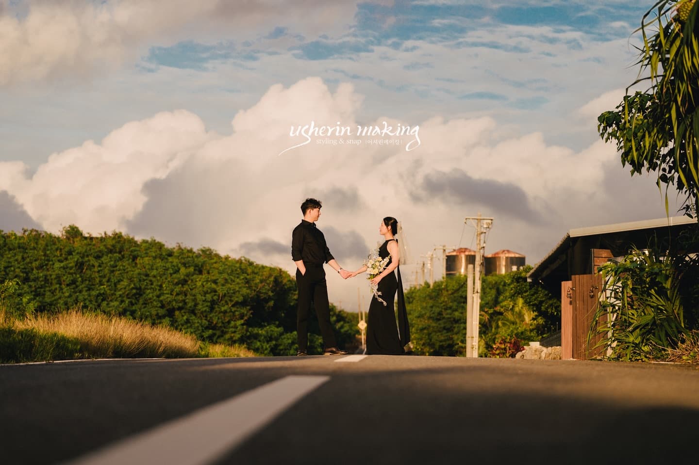 Bride and groom in black holding hands on a quiet road under a cloud-filled sky in Miyakojima