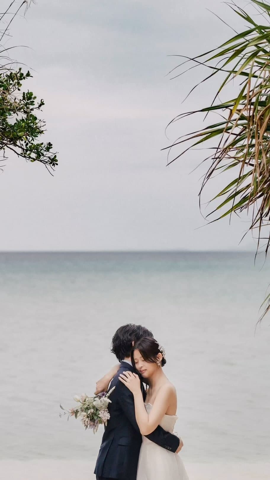 曇り空の静かな海辺でそっと抱き合う新郎新婦、沖縄のビーチフォト
