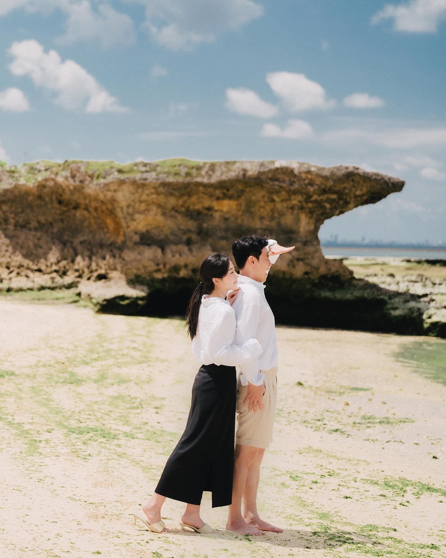 A couple strolls along the sand past a large rock under Okinawa's blue sky