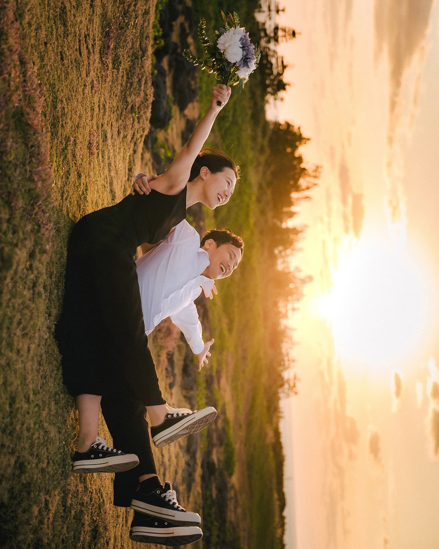 Woman on her partner's back raising a bouquet toward the sunset glow in Okinawa