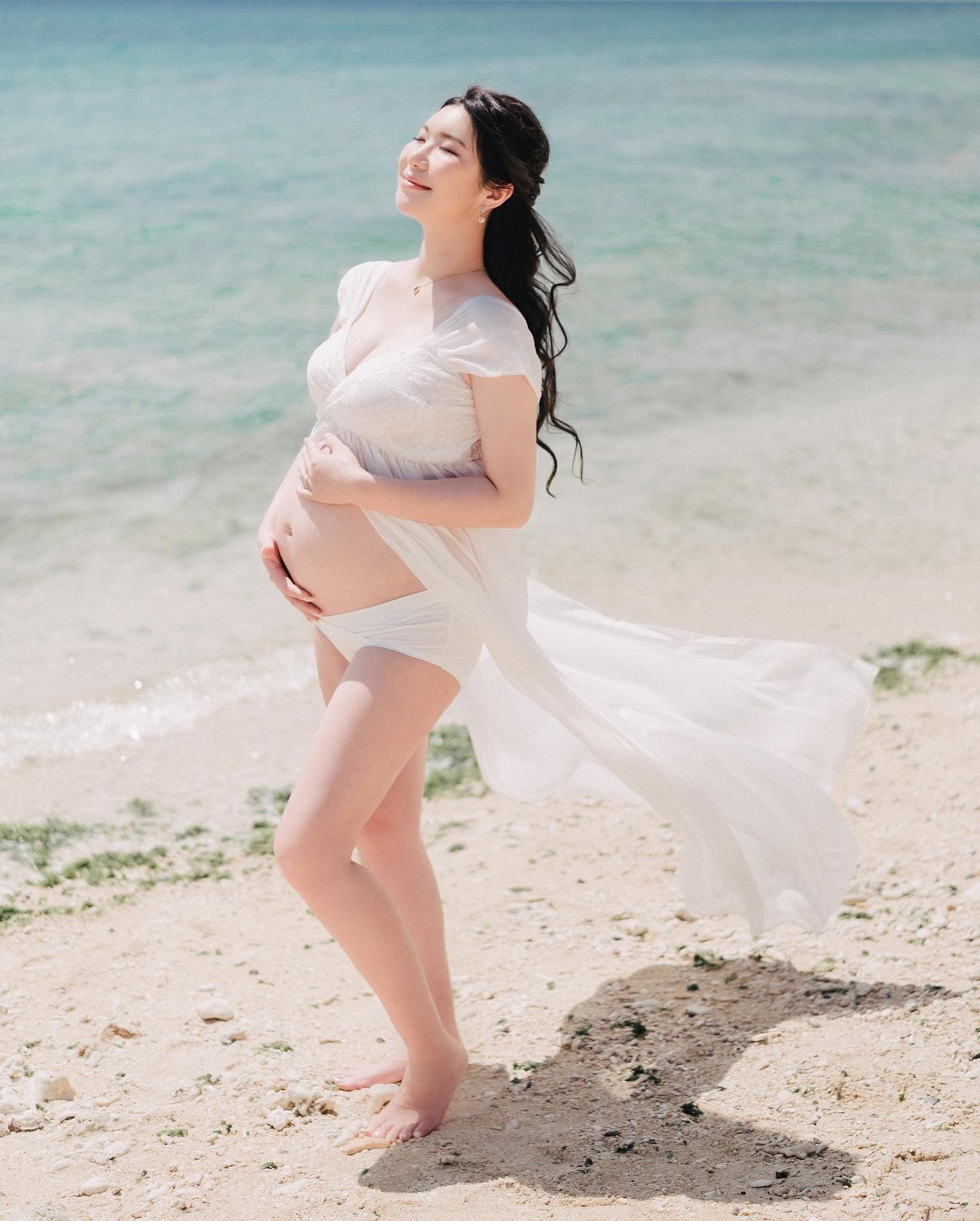 Expectant mother in a flowing white dress cradling her belly on an Okinawa beach