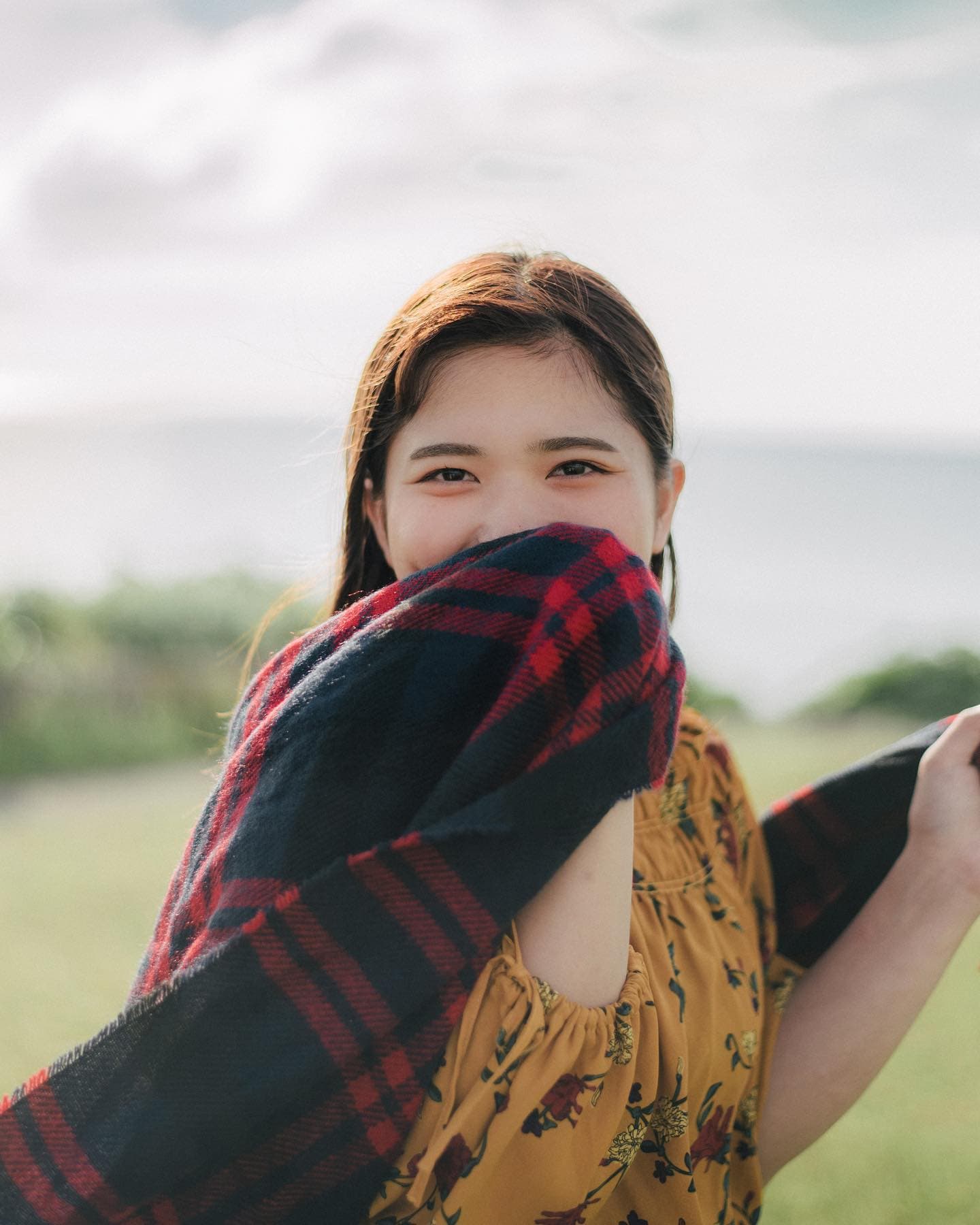 Woman smiling behind a plaid scarf on a grassy seaside hill in Okinawa