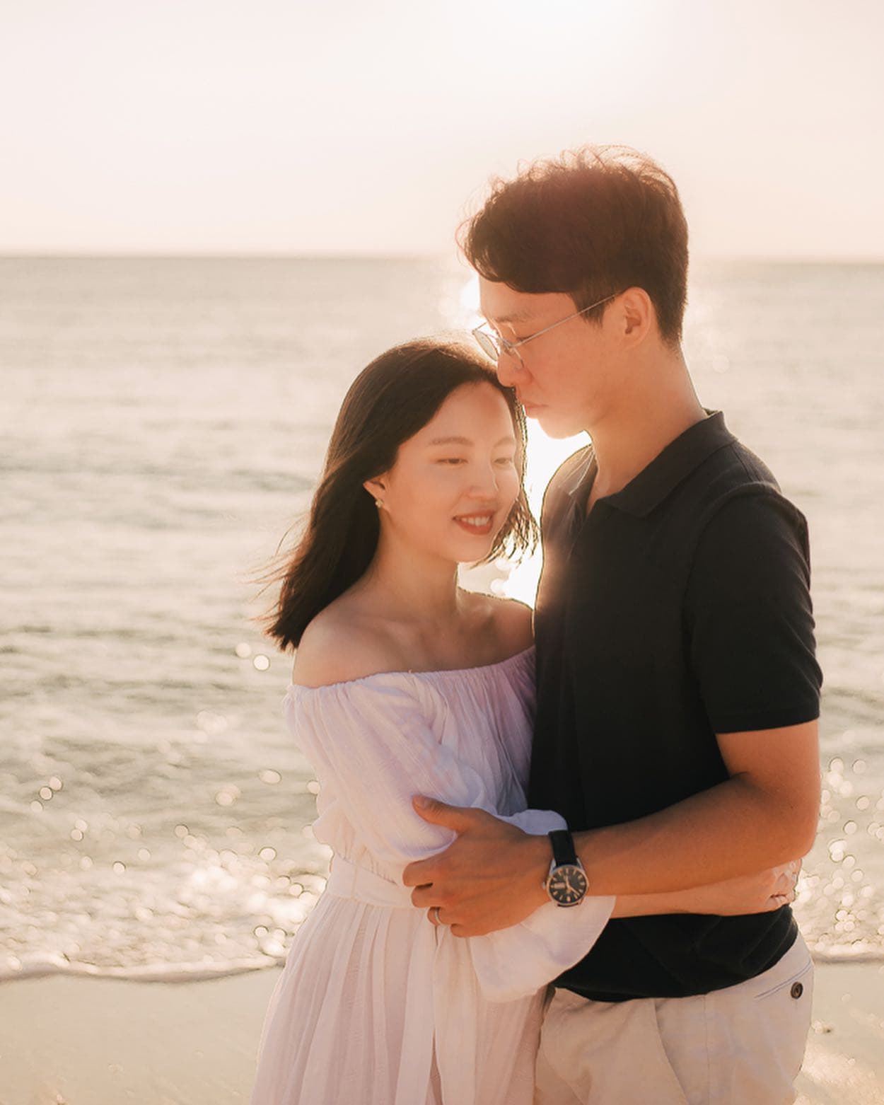 환한 오키나와 해변에서 서로 껴안은 흰 원피스의 여성과 남색 셔츠의 남성