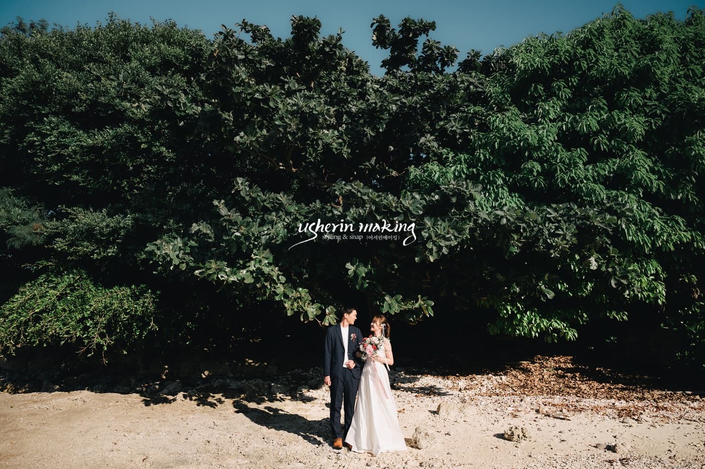 Couple in wedding attire standing on the sand before deep green trees in Okinawa