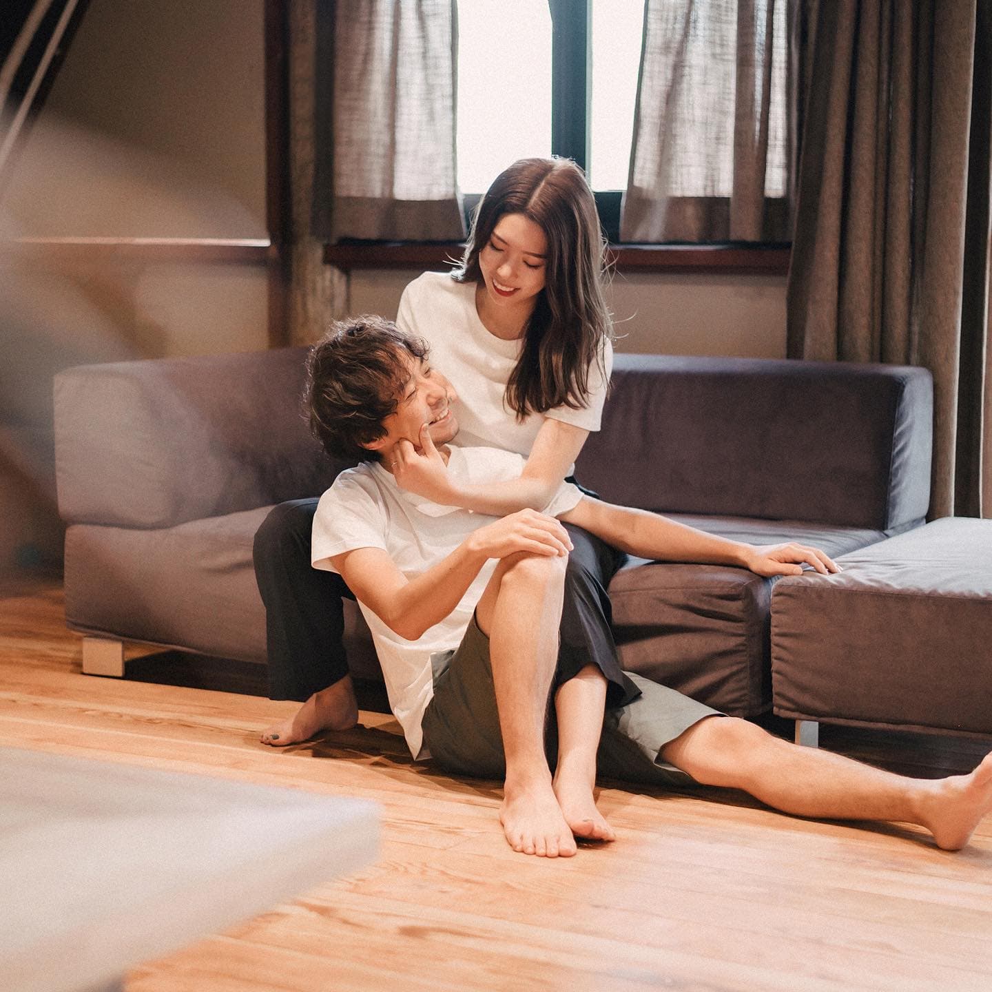 Couple in casual clothes smiling at each other beside a gray sofa indoors
