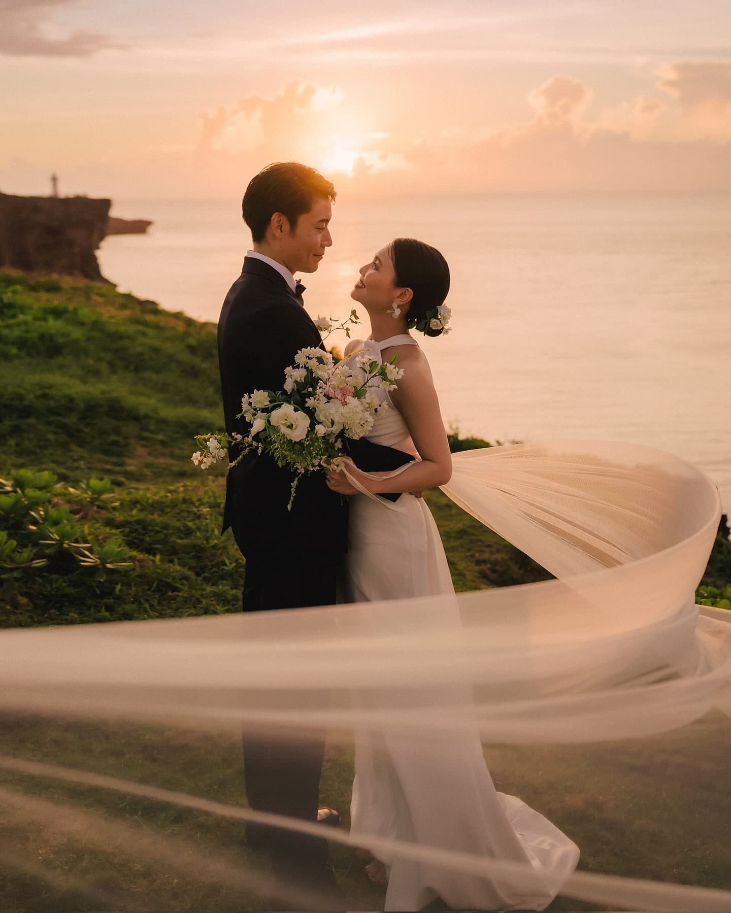 風に舞うベールと白い花束、夕日の海辺で見つめ合う新郎新婦、沖縄