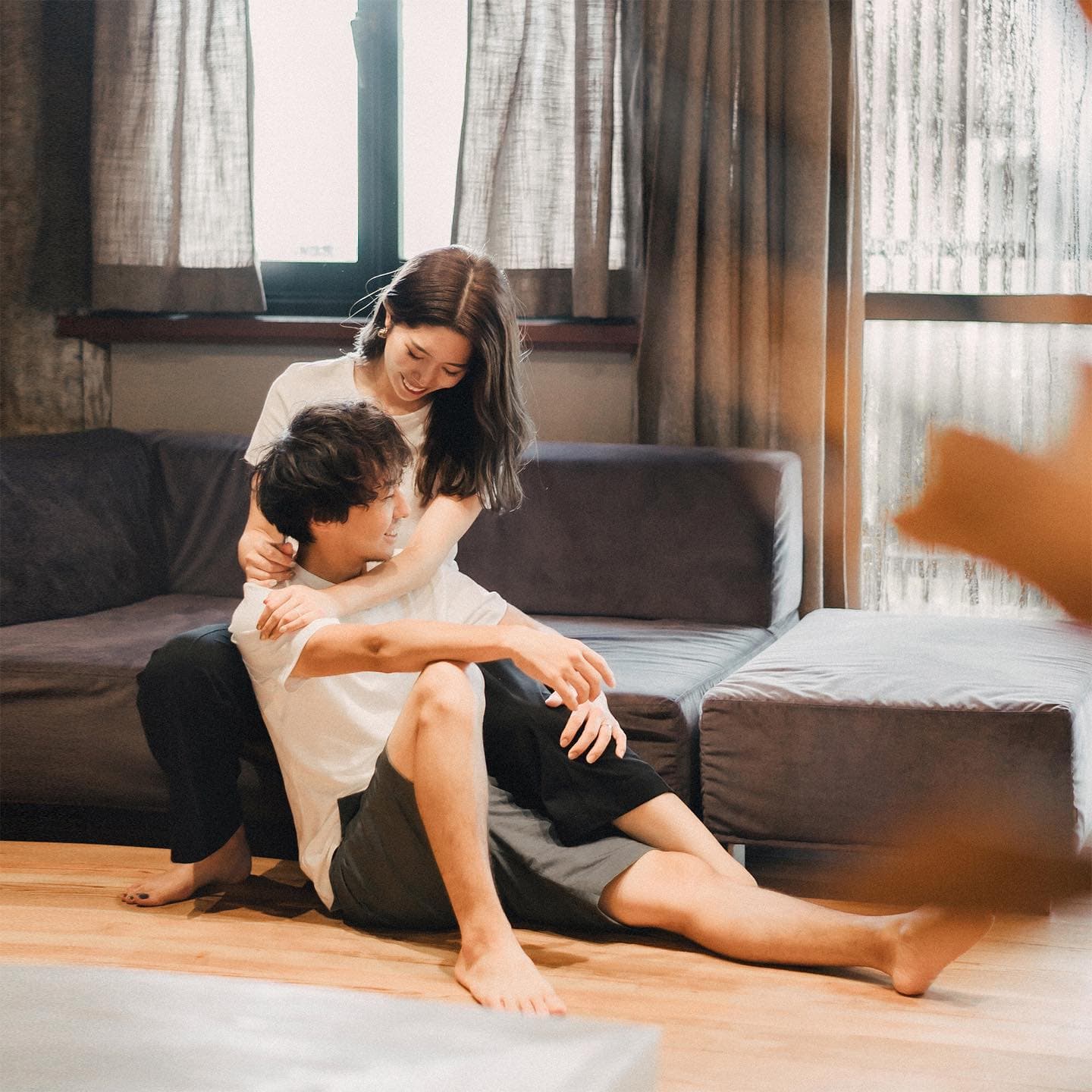 Woman hugging her partner from behind by a sofa near the window