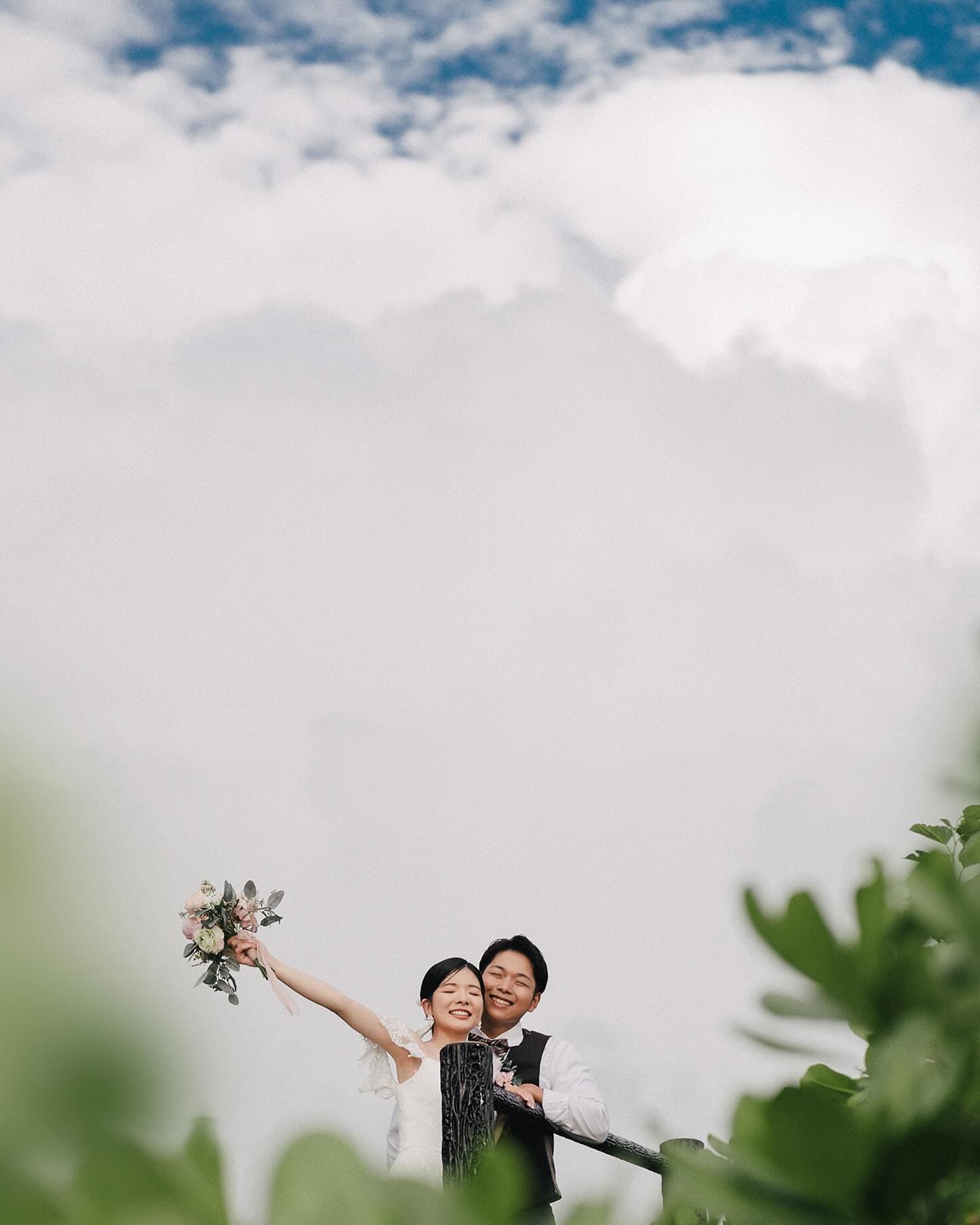 Couple laughing, bouquet raised, before towering clouds in Okinawa