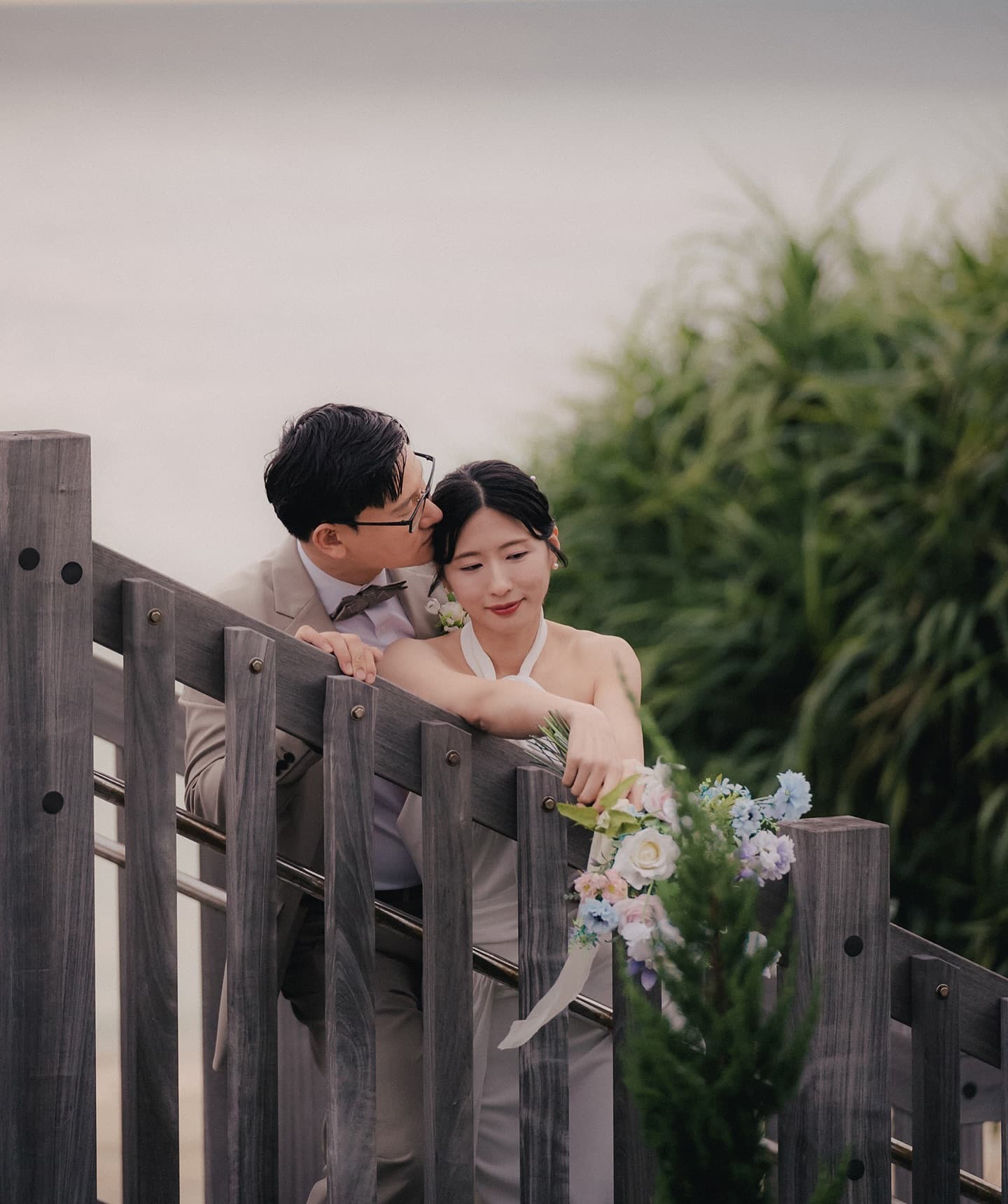 Groom kissing the bride's head at a wooden seaside fence at dusk in Okinawa