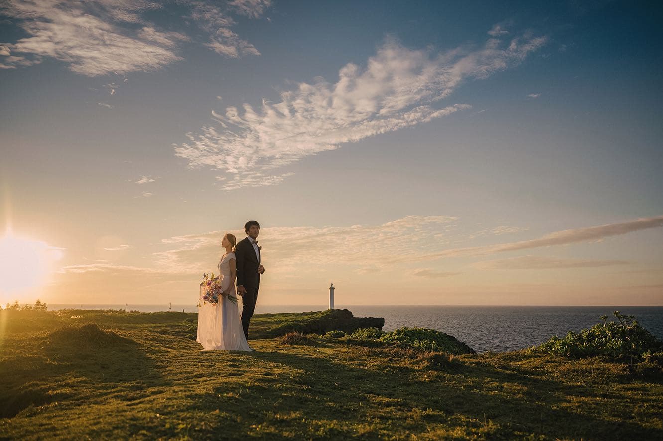 Bride and groom on a grassy cape at sunset, a lighthouse on the horizon in Okinawa