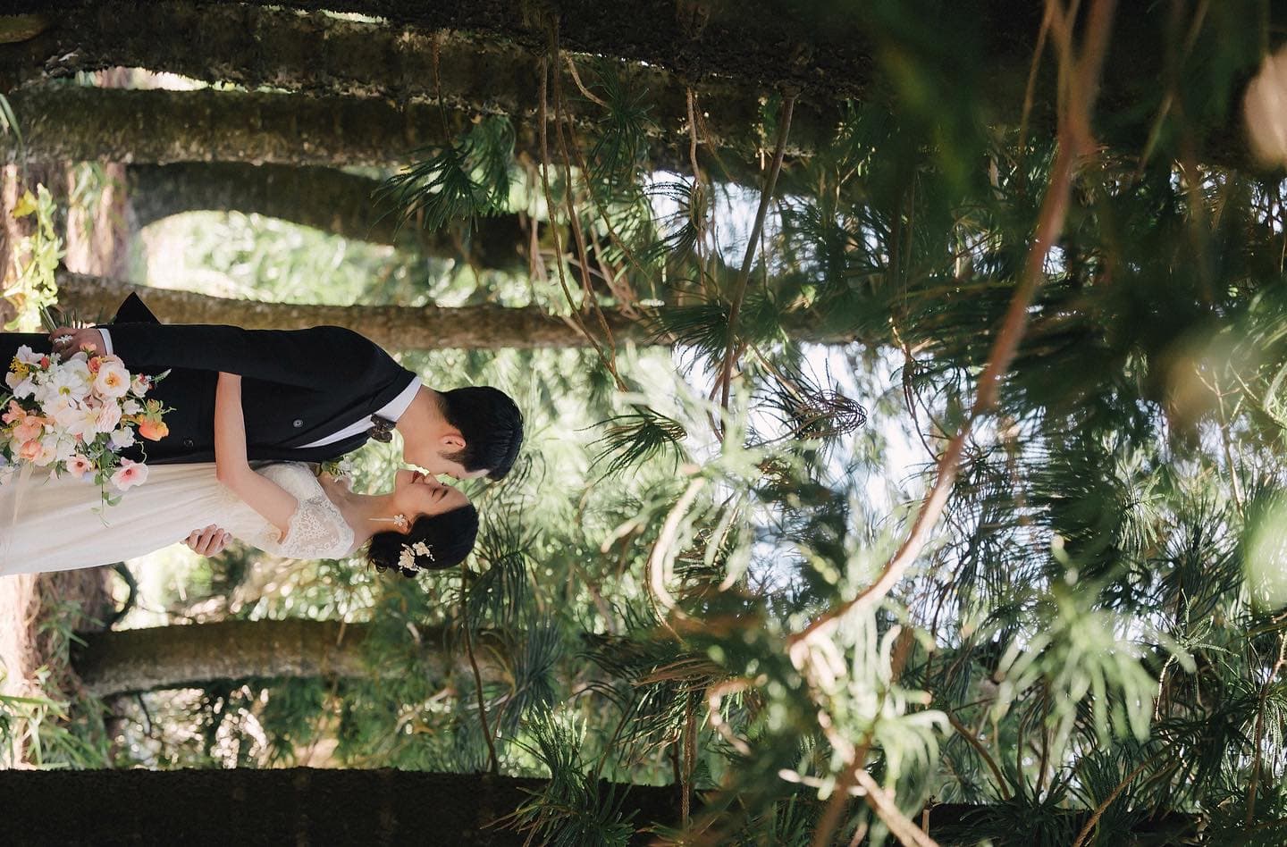 Couple kissing beneath tall pine branches, bouquet in hand, Okinawa