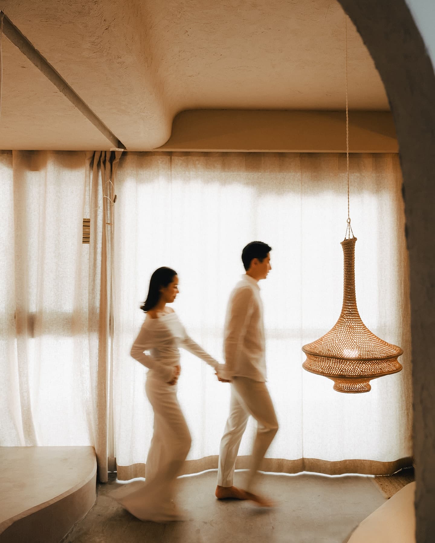 A couple in white walks hand in hand through a softly lit studio