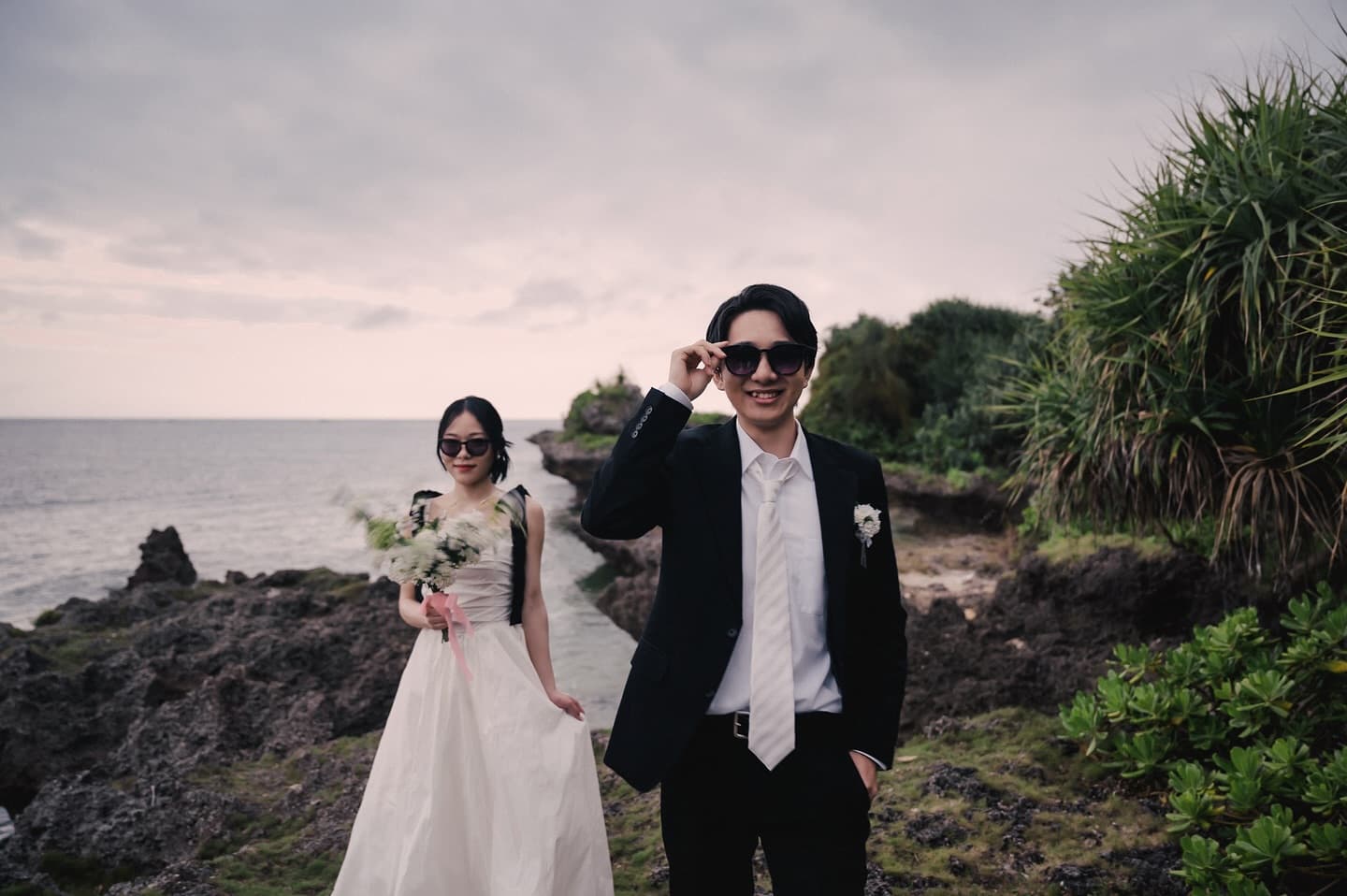 Bride and groom in sunglasses posing on a rocky coast under a cloudy sky in Okinawa