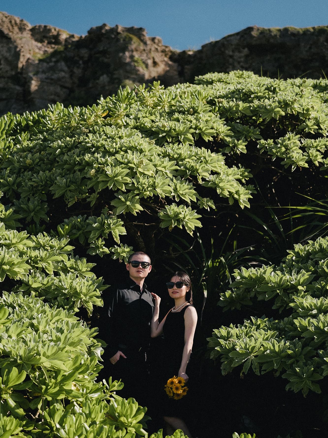 Couple in black standing among lush green shrubs with sunflowers on Miyako Island, Okinawa