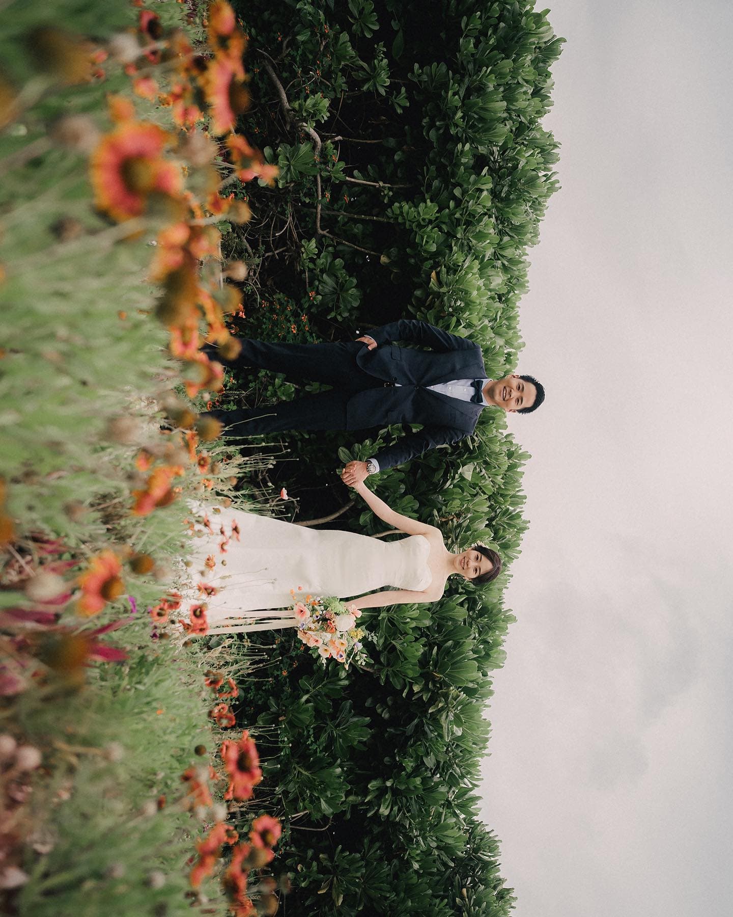 Bride and groom holding hands among green shrubs dotted with orange wildflowers