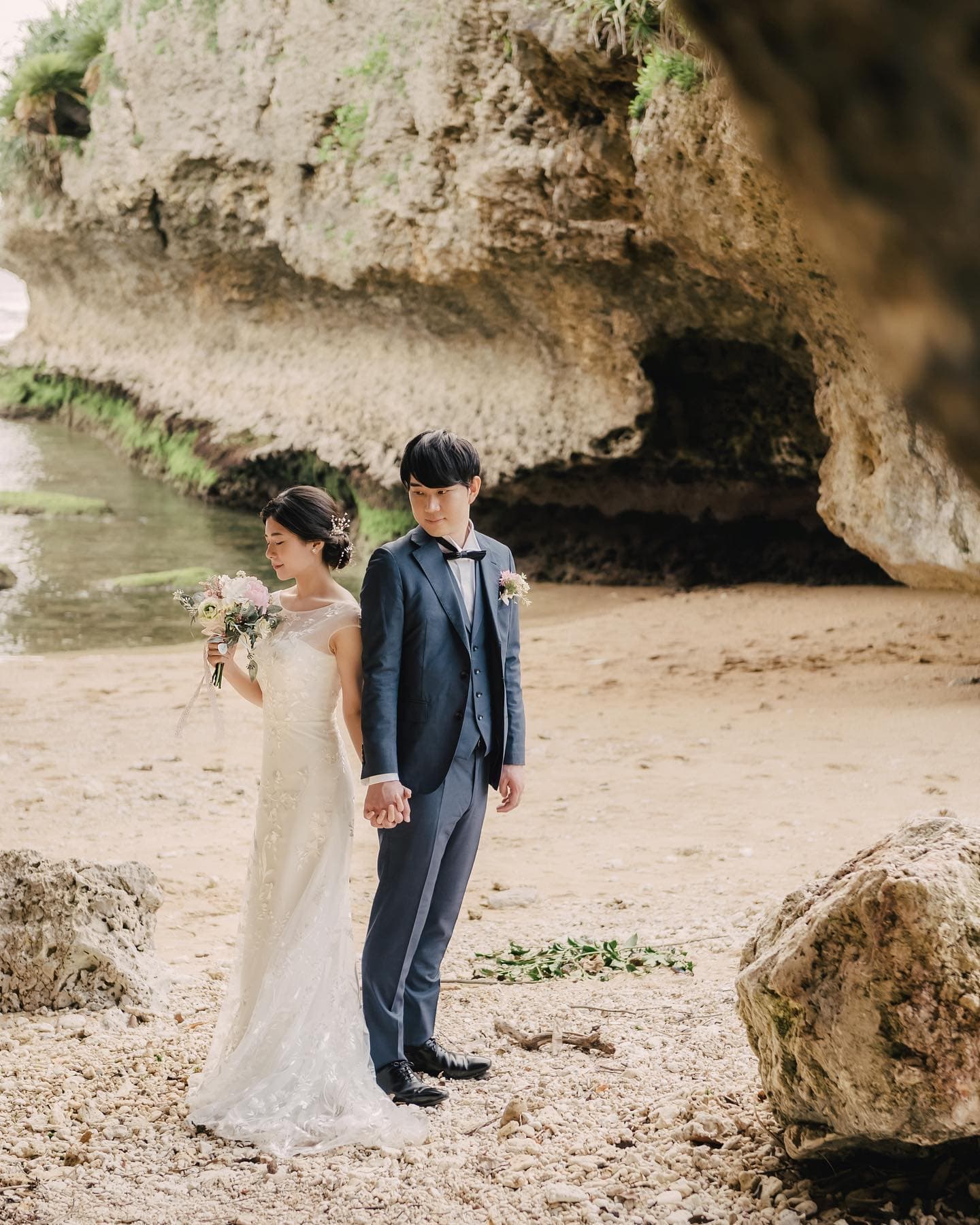 岩の洞窟のあるビーチで手をつなぐ新郎新婦。沖縄ロケーション。