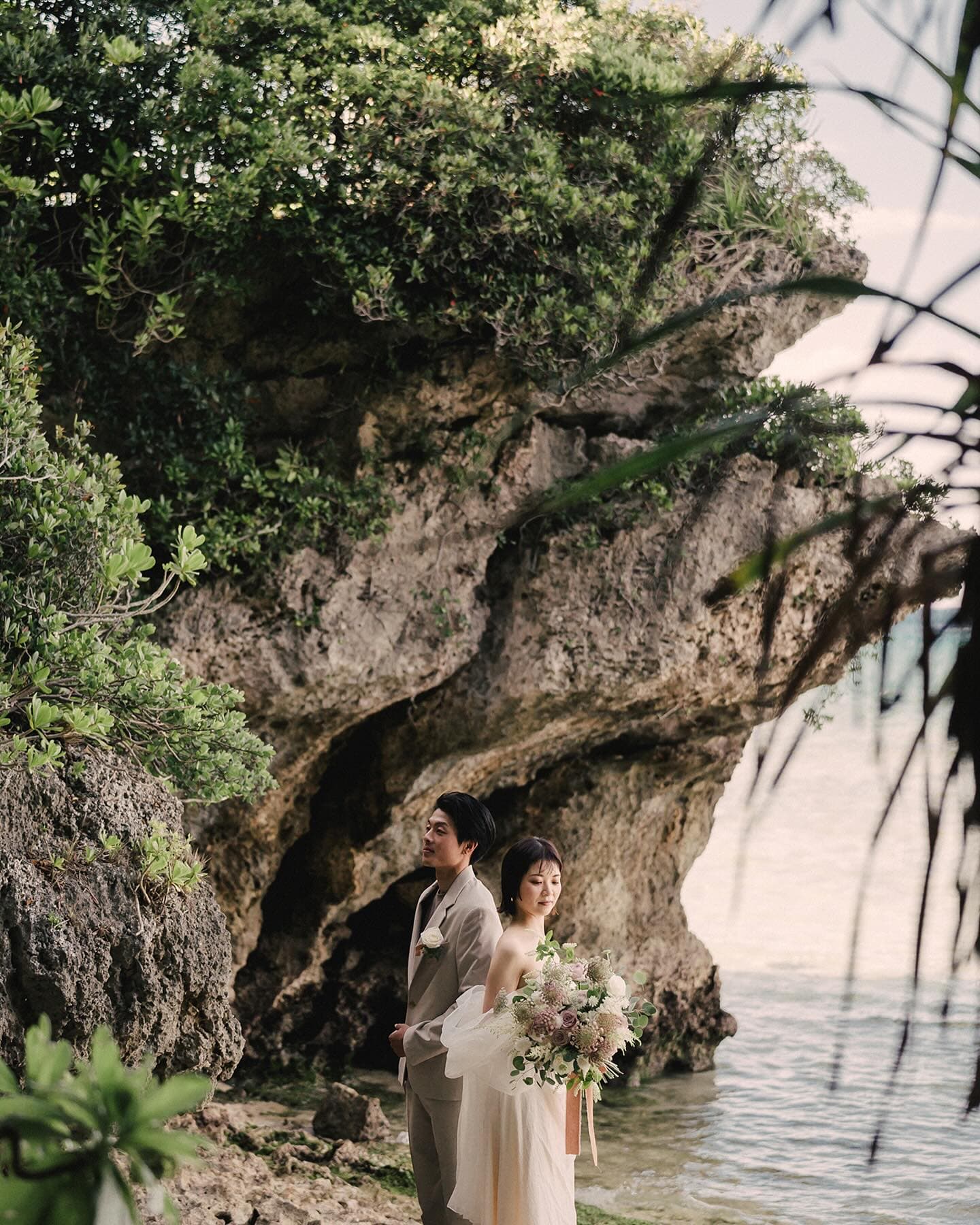 Bride and groom standing beneath a huge green-covered rock formation by the Okinawa sea