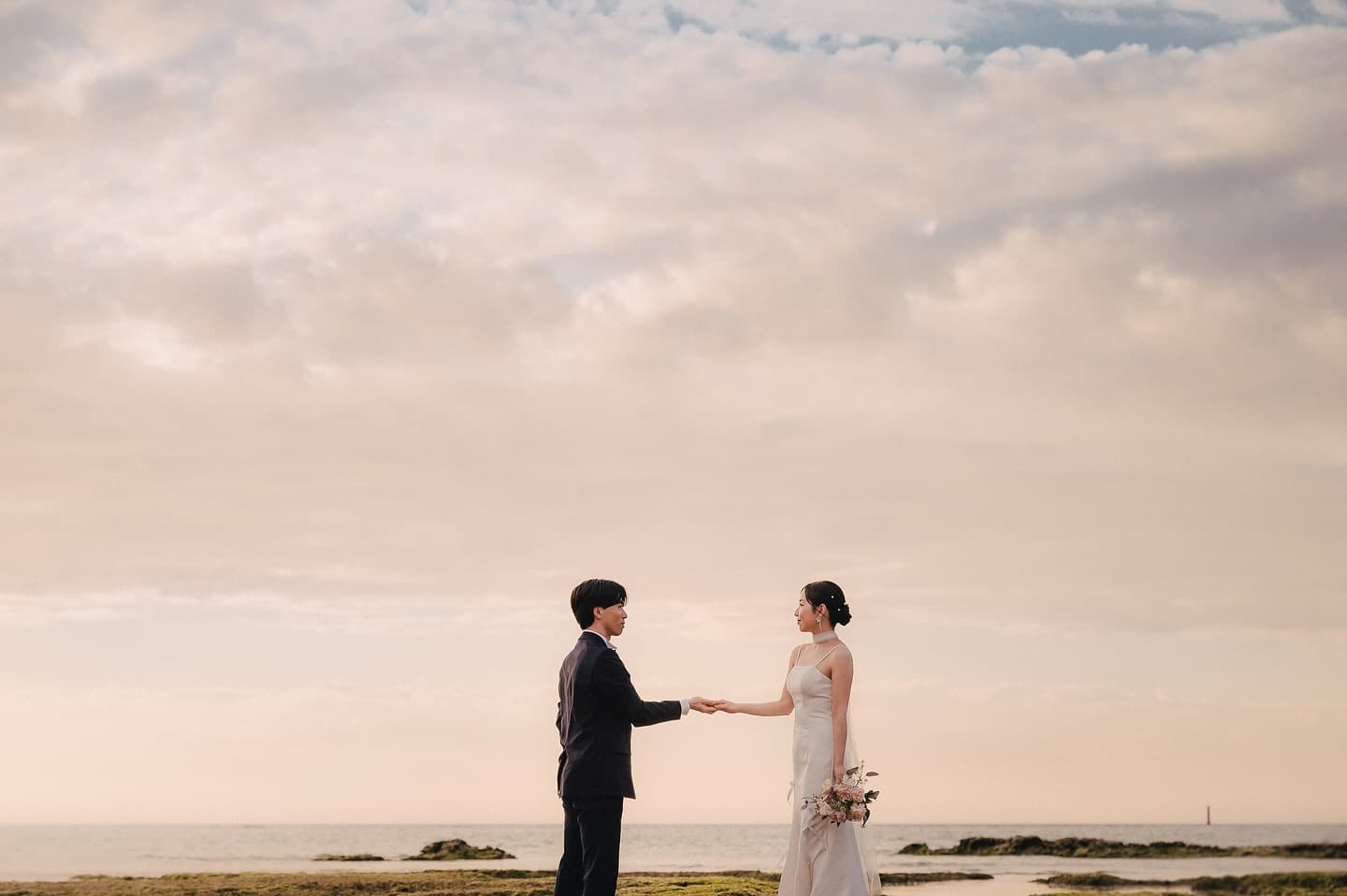 Bride and groom holding both hands by the sea under a soft pink sunset sky in Okinawa
