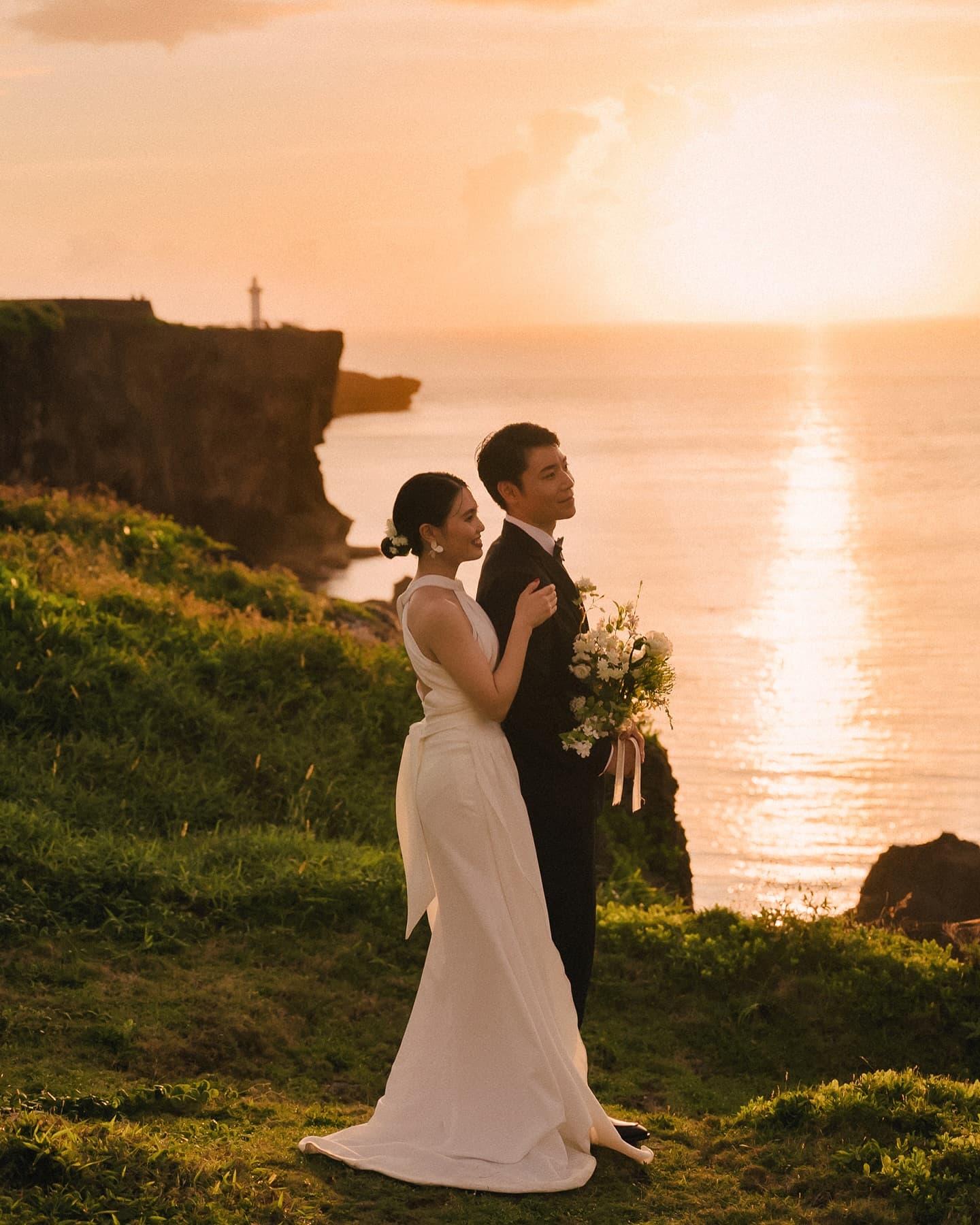 Bride and groom on a grassy Okinawa clifftop above the sunset sea