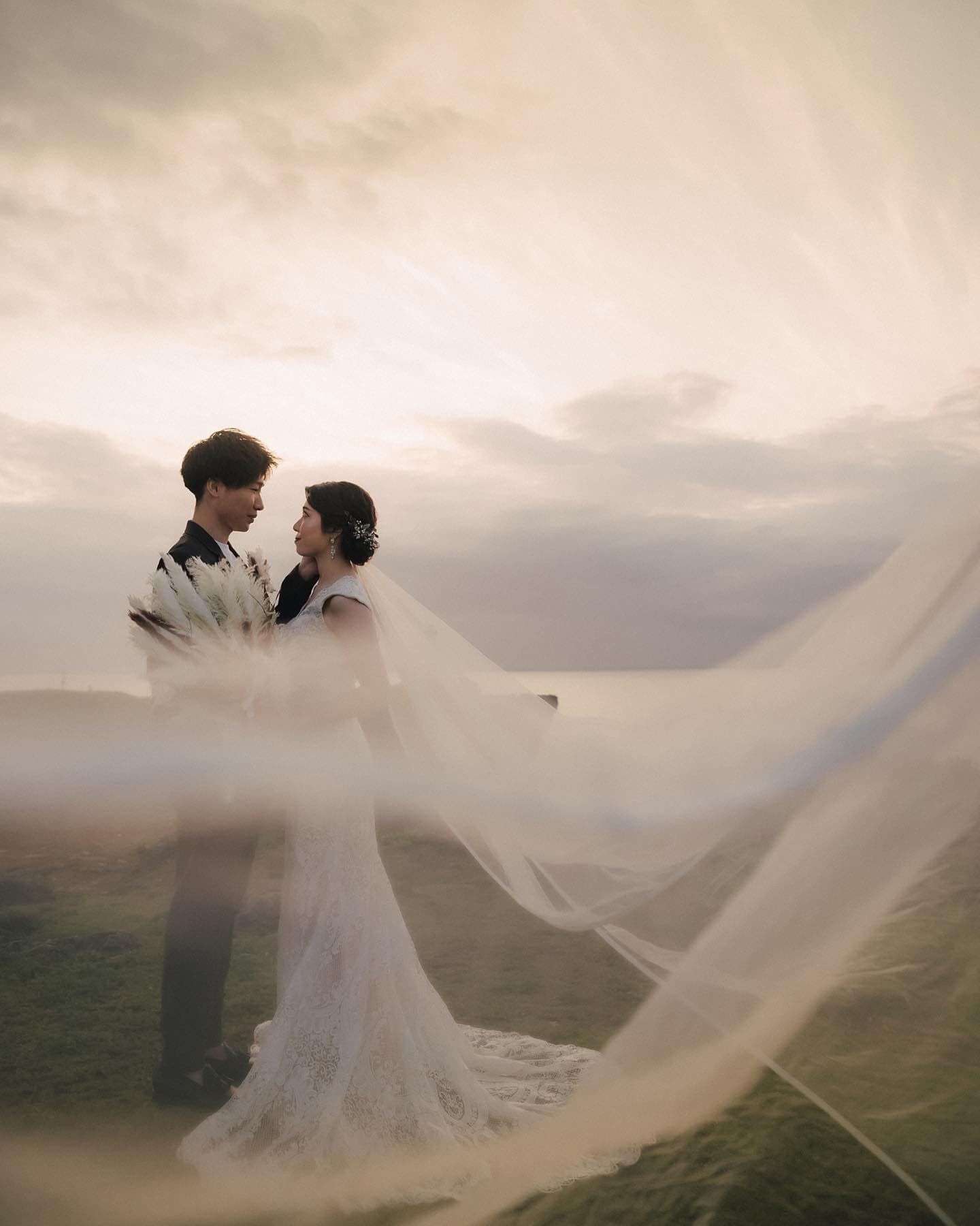 A long veil sweeps through the air as a couple face each other under cloudy Okinawa skies