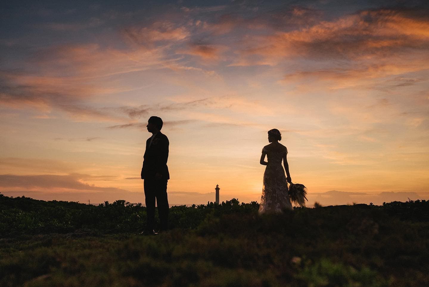 Two silhouettes stand on a twilight hill in Okinawa, a lighthouse between them