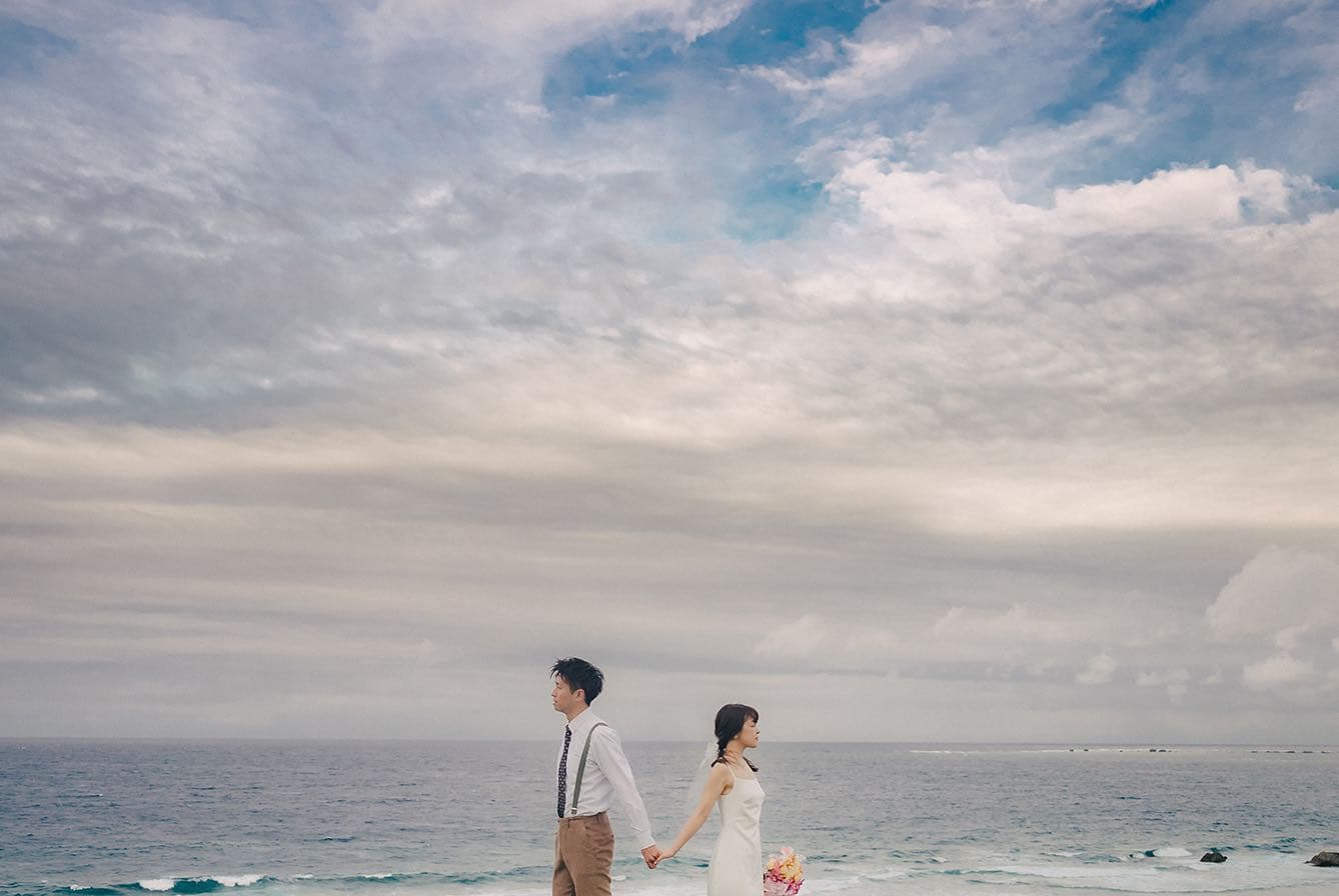 Bride and groom holding hands on a Miyakojima beach beneath a wide cloudy sky
