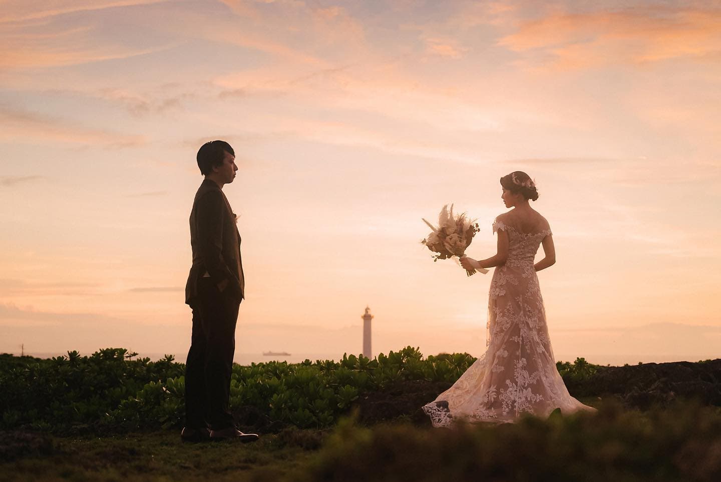 Bride and groom face each other at dusk on an Okinawa cape, a lighthouse in the distance