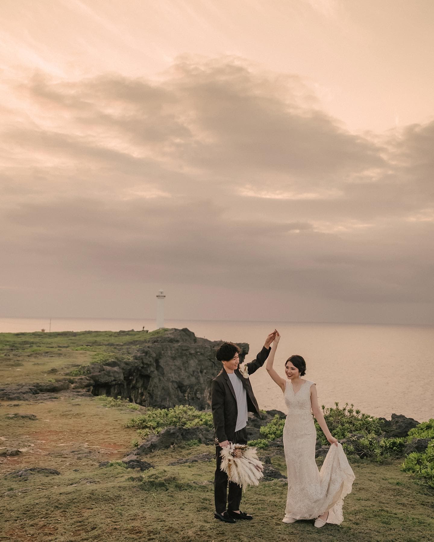 A couple join hands on an Okinawa cape at dusk beneath a soft cloudy sky