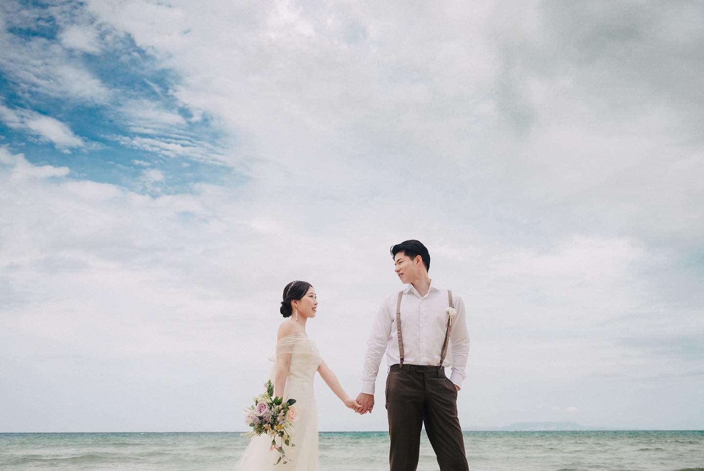 Bride and groom joining hands on an Okinawa beach beneath a wide summer sky