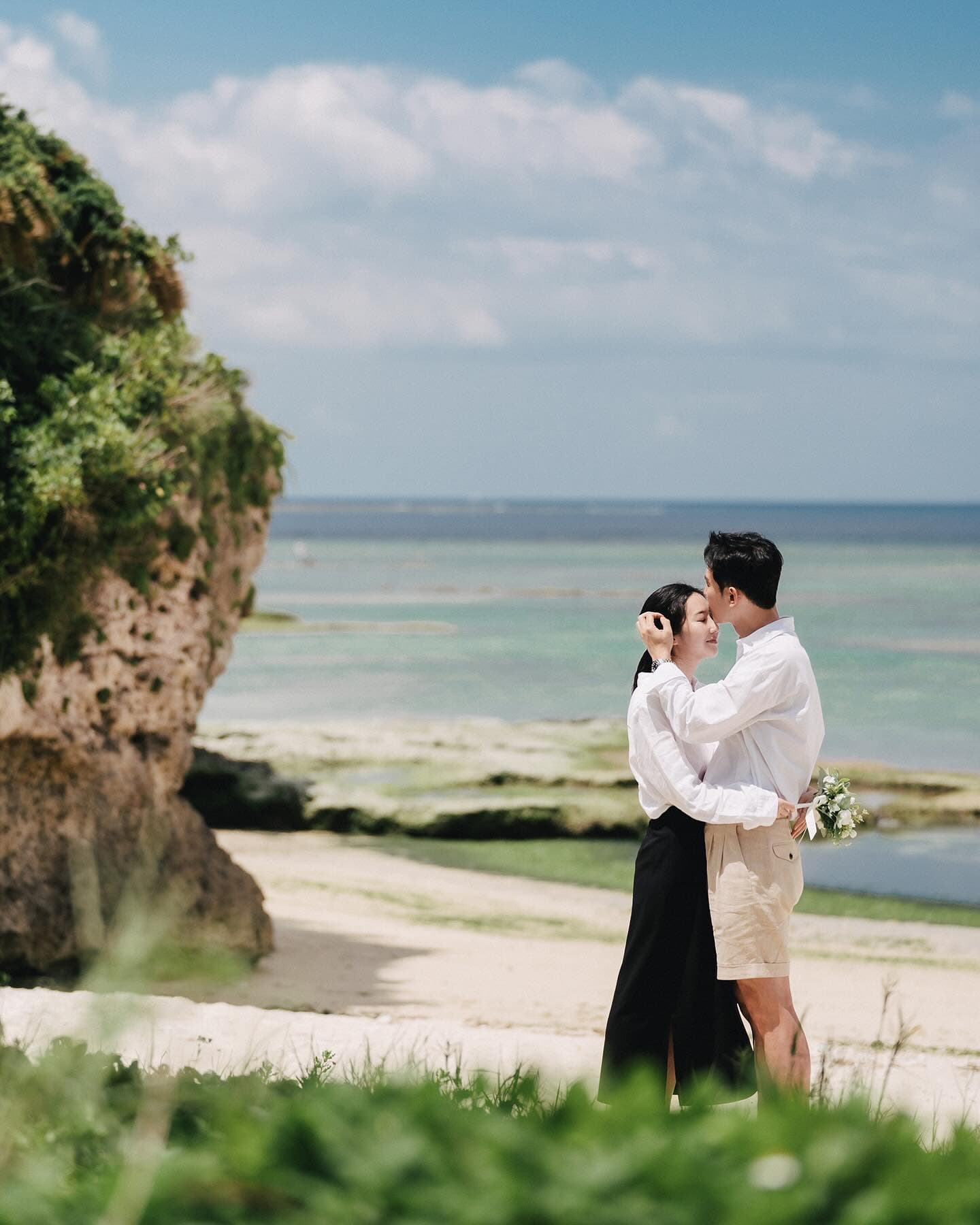 A couple touches foreheads by rocky cliffs and the emerald sea in Okinawa