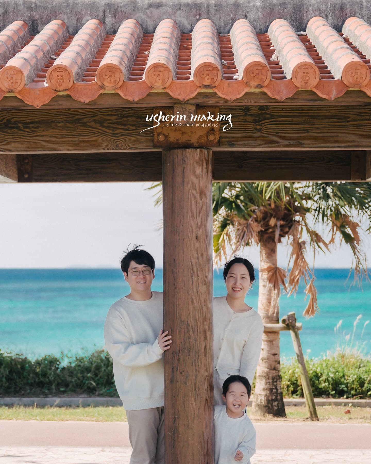 Family smiling around a pillar under a red-tiled pavilion by the emerald sea in Okinawa