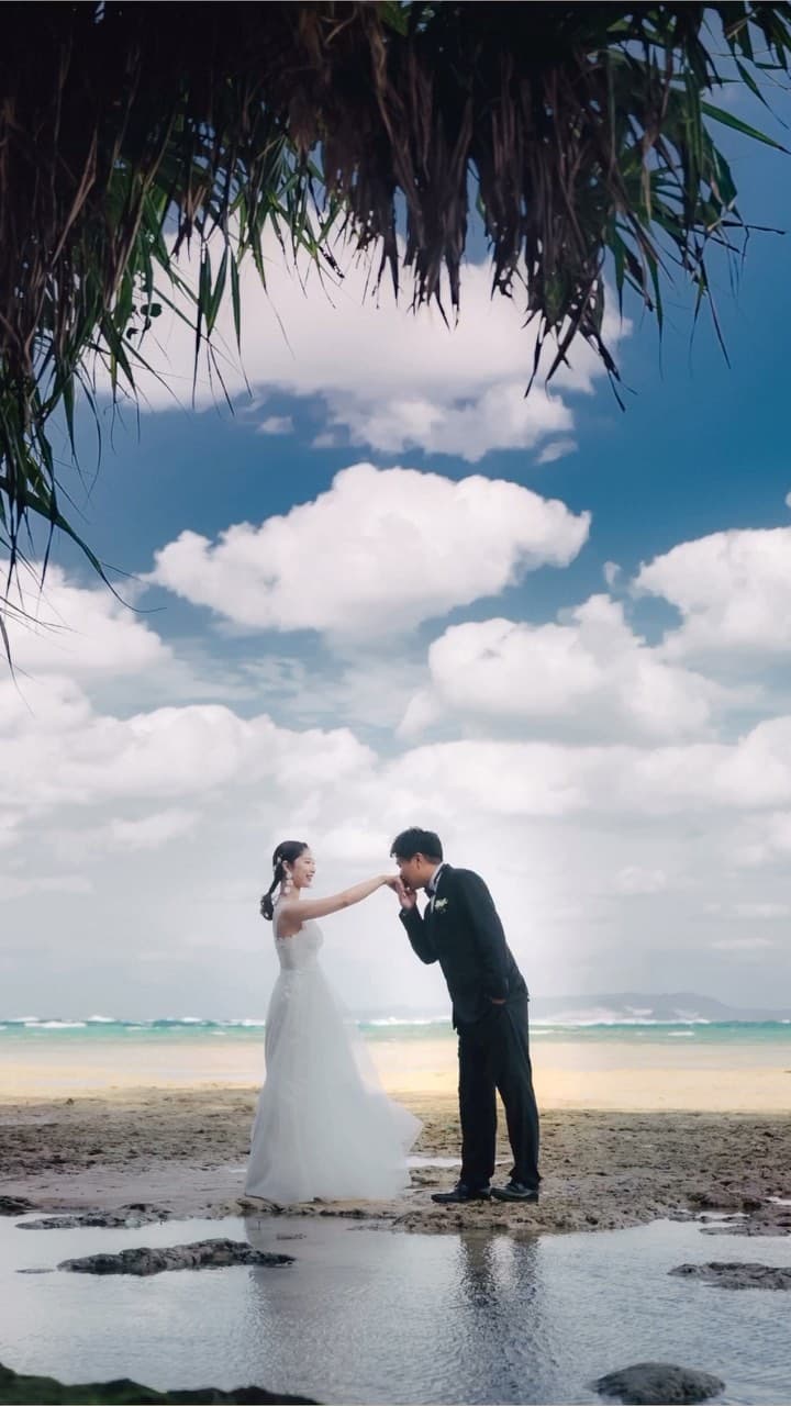 Groom kisses the bride's hand on an Okinawa beach beneath towering summer clouds