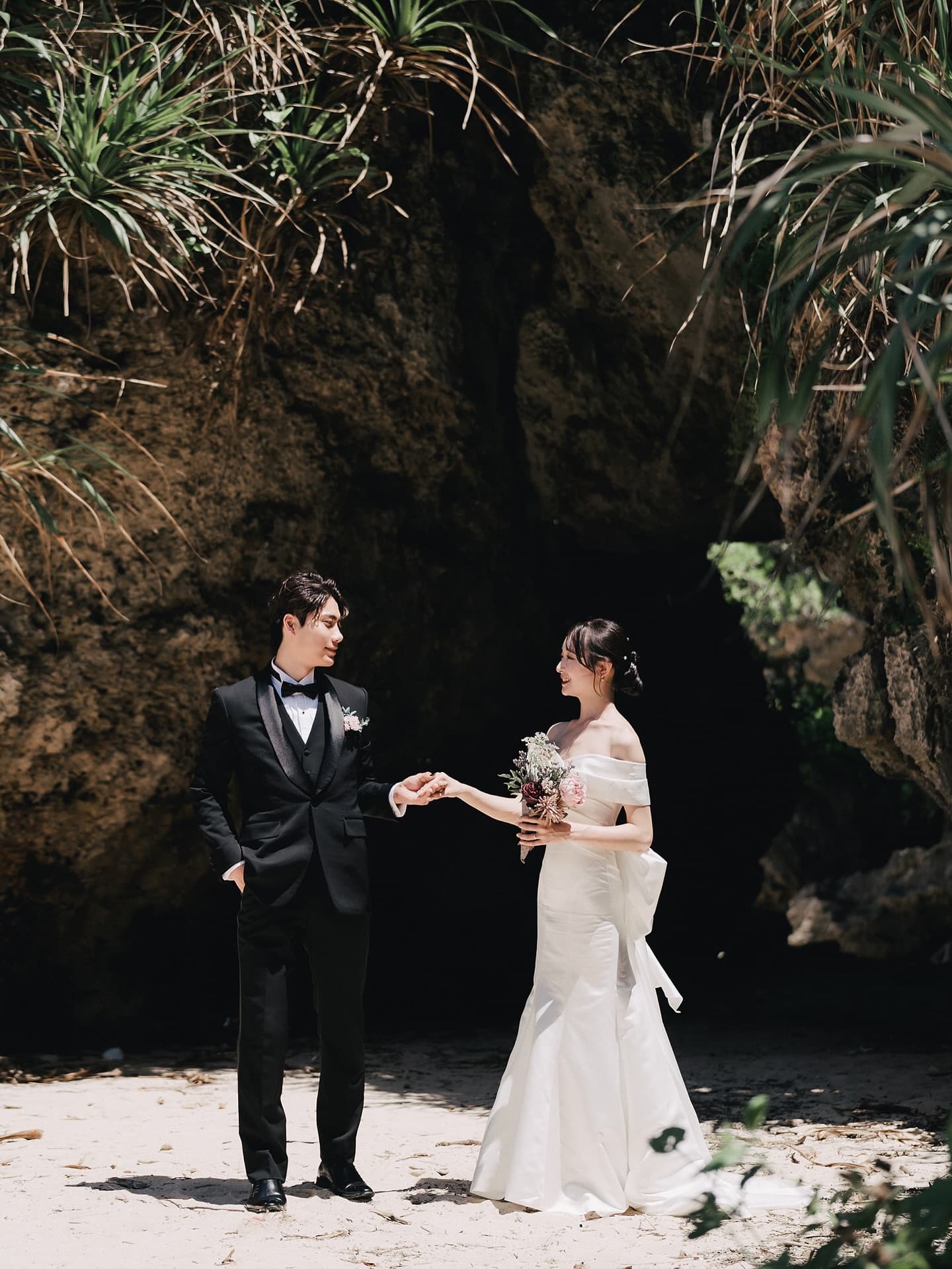 Groom in a tuxedo taking the bride's hand before a plant-draped rock cave in Okinawa