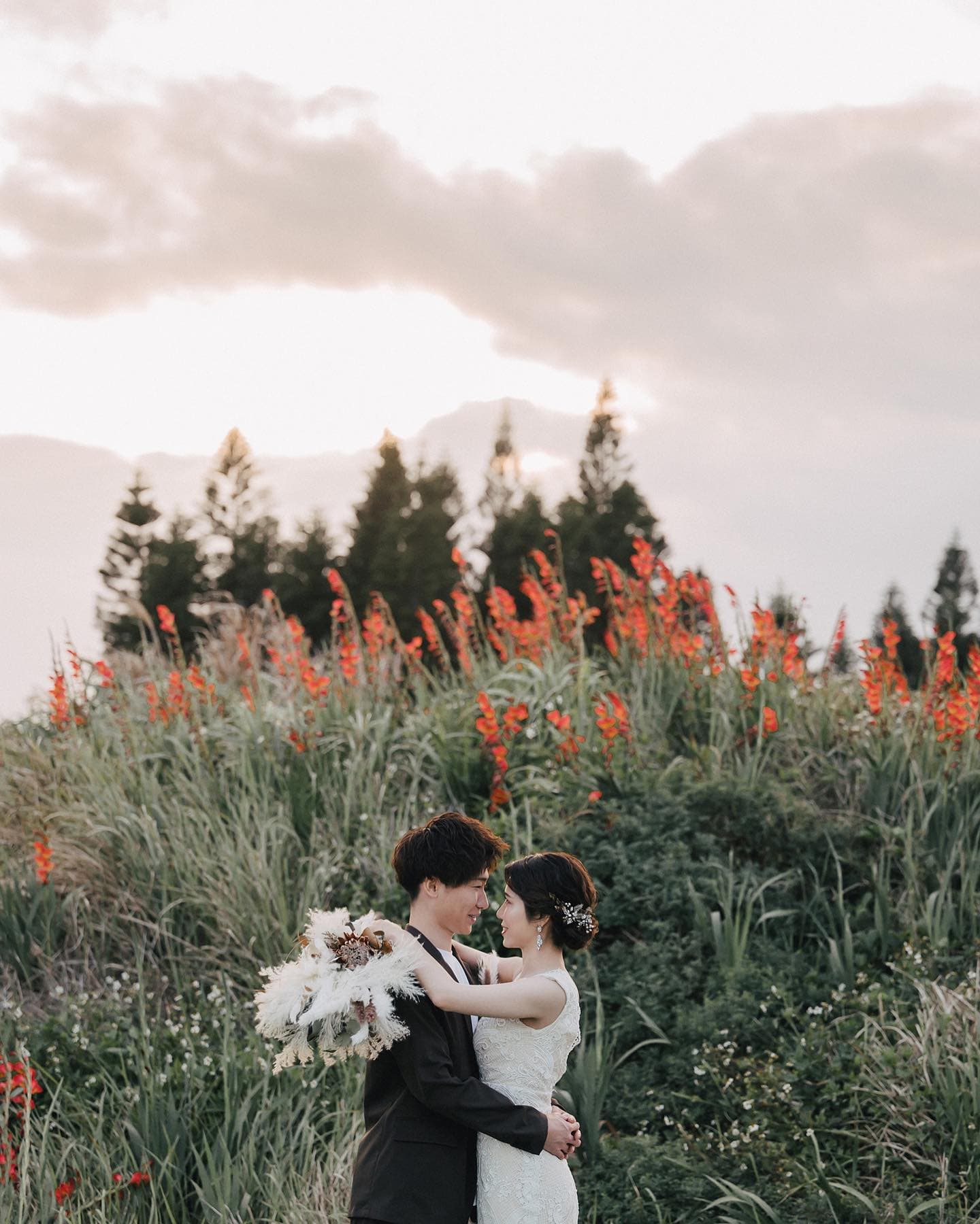 Bride and groom embrace in a grassy field of red blooms under an overcast sky in Okinawa