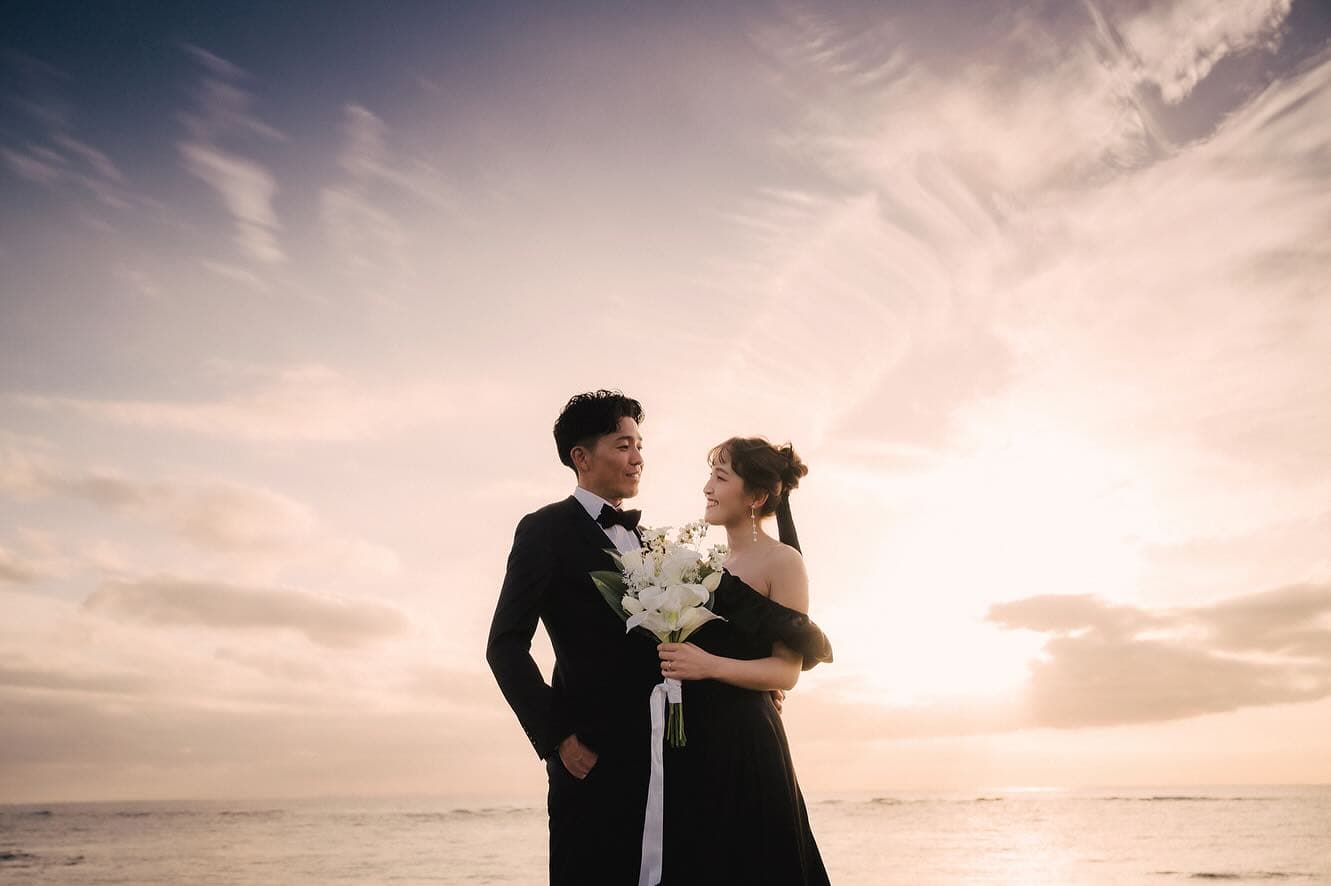 Couple in black attire with a white bouquet under a pastel sunset sky, Okinawa