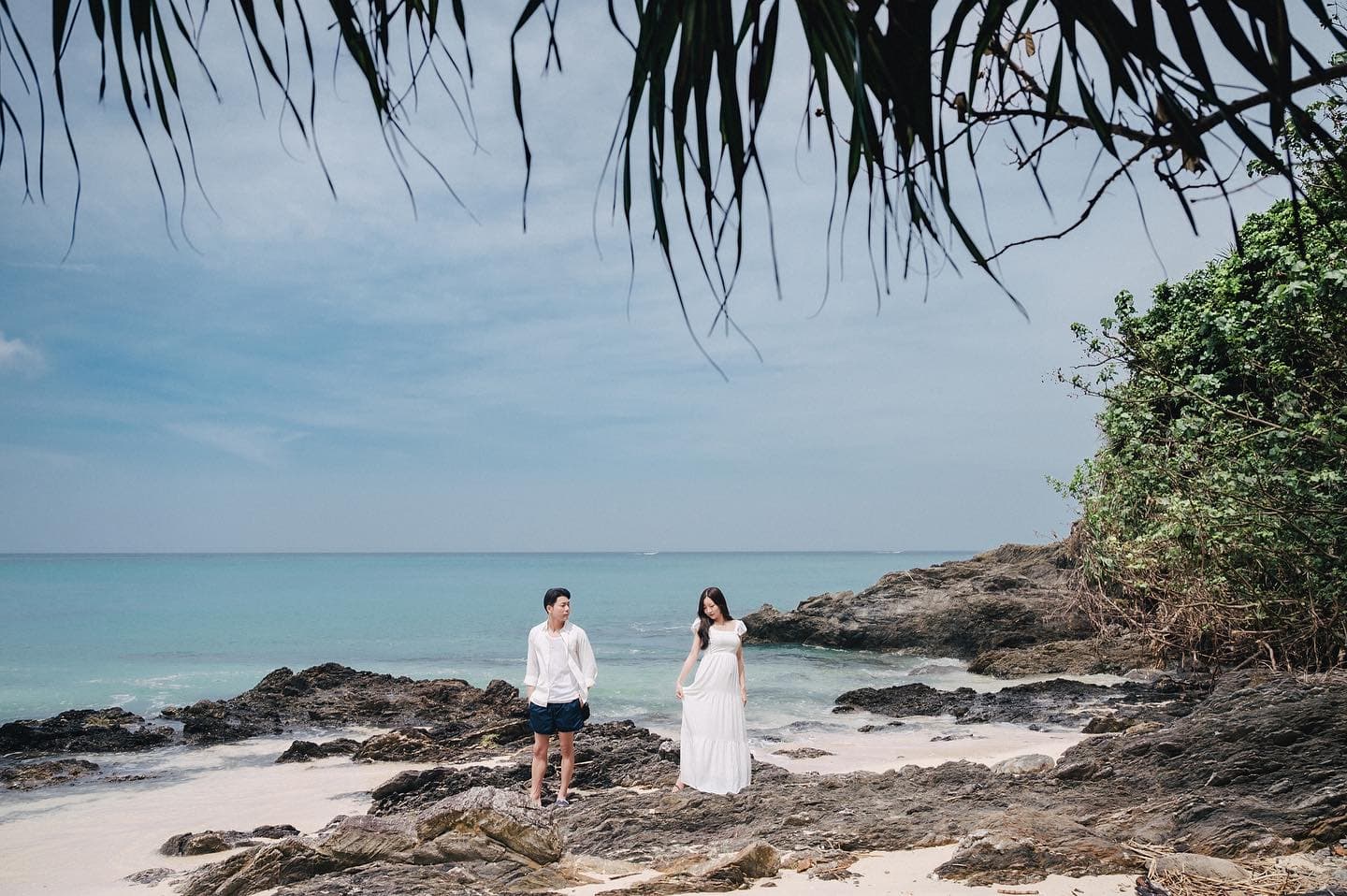 Couple in white on a rocky beach framed by palm leaves, Okinawa