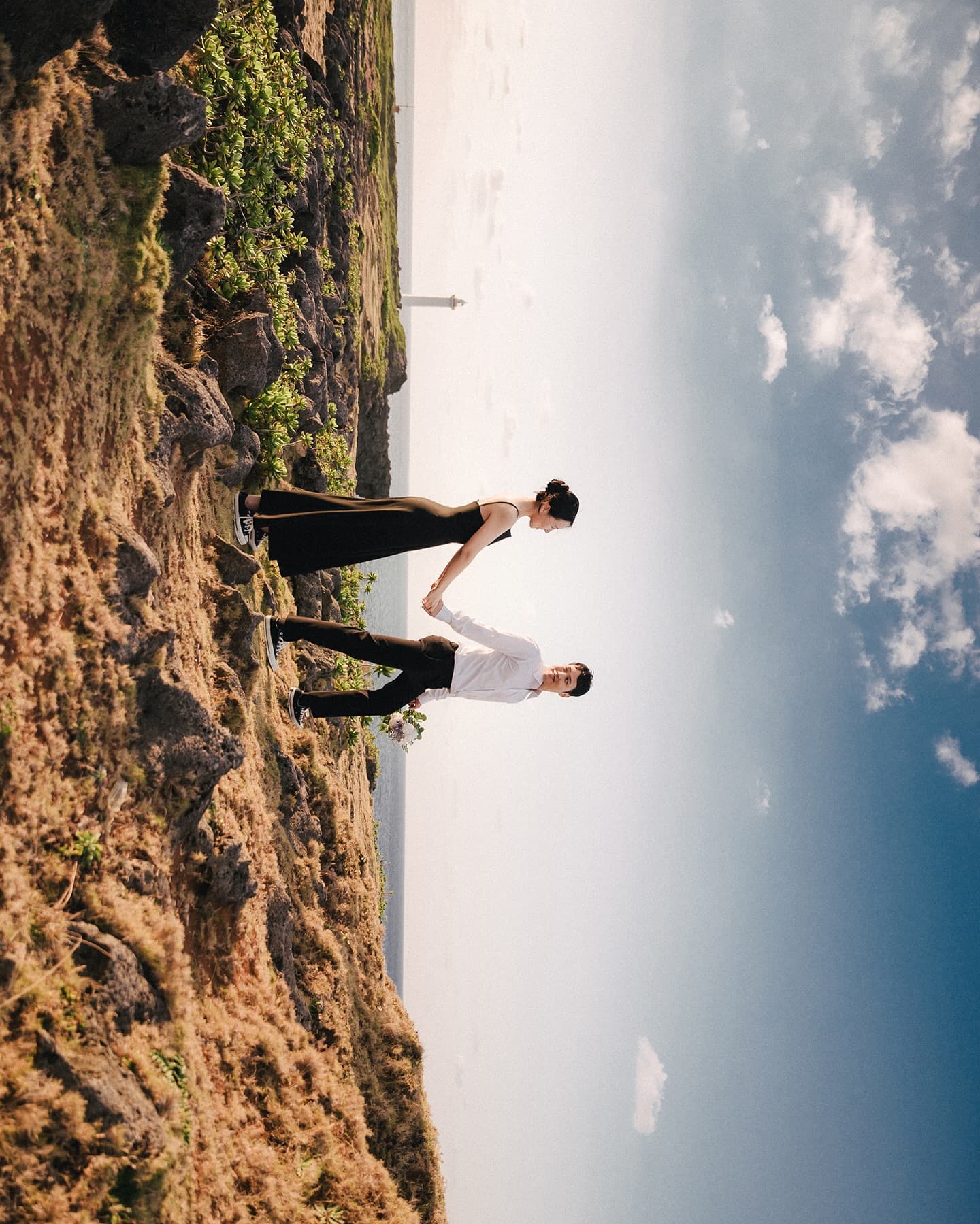 Couple holding hands on a rocky hill under a bright blue sky in Okinawa