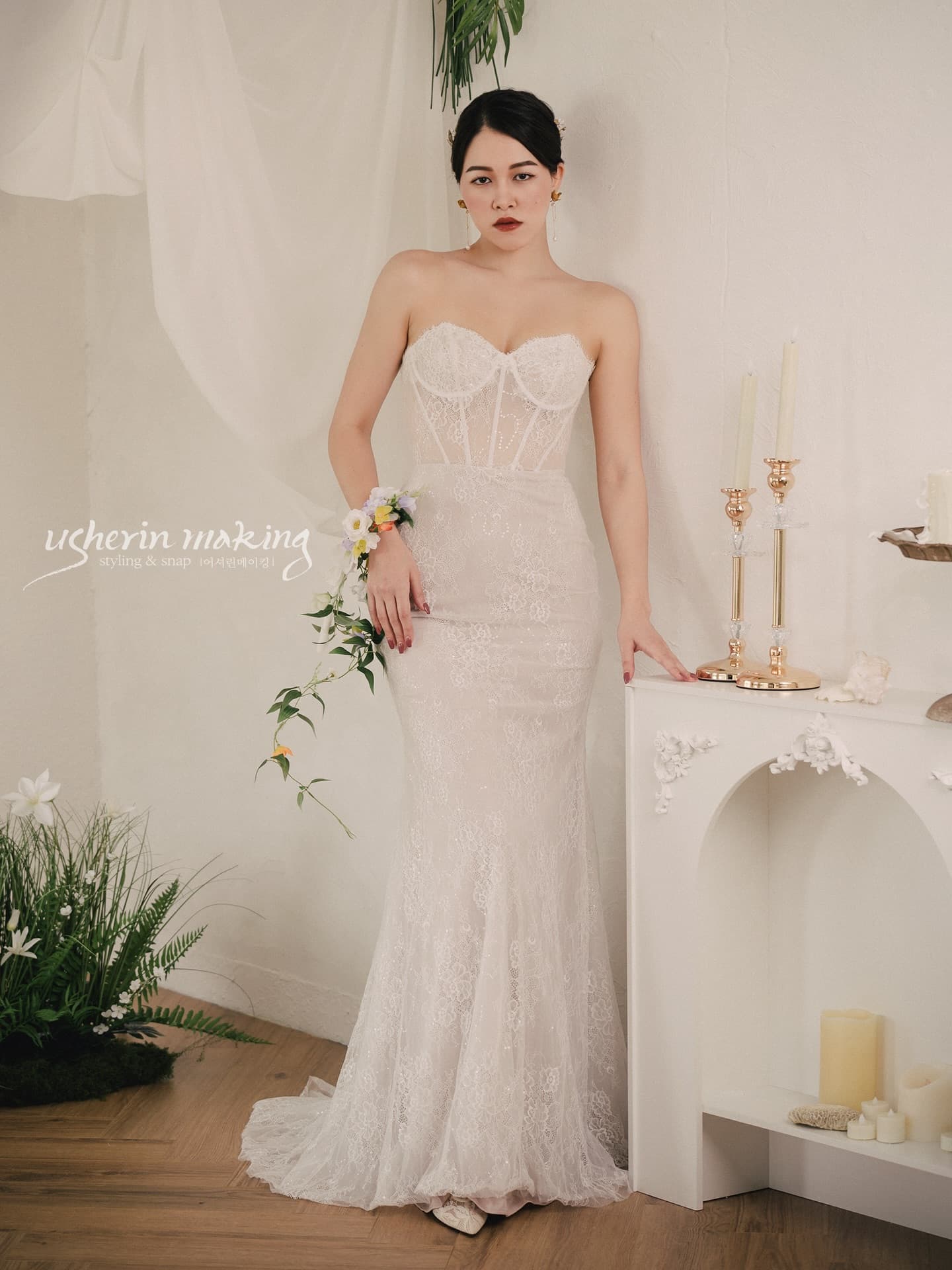 A bride in a lace mermaid dress stands by a white mantel and candlesticks