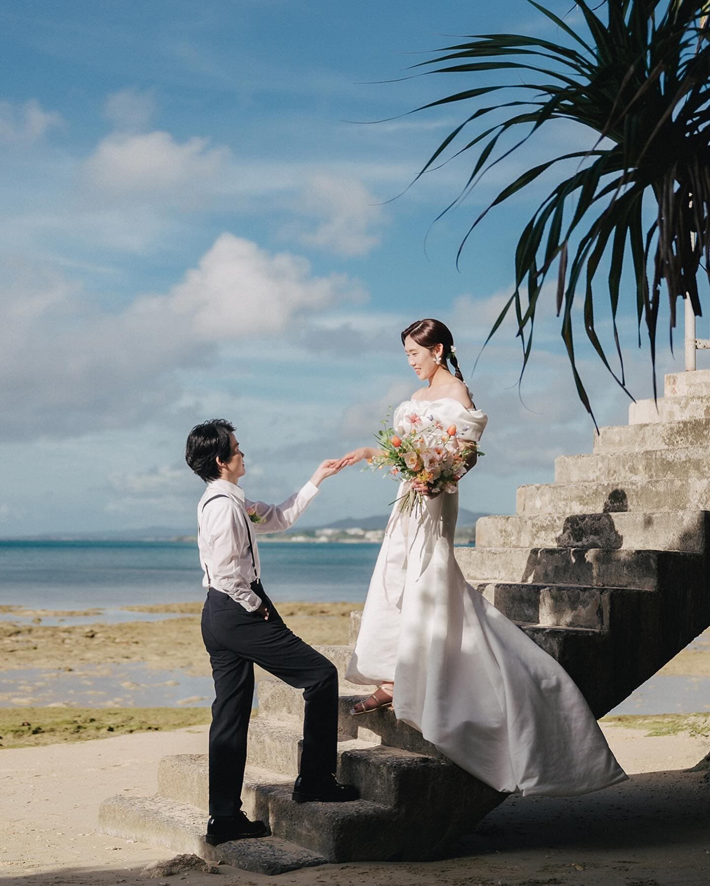 Groom offering his hand to the bride coming down stone steps by the sea in Okinawa