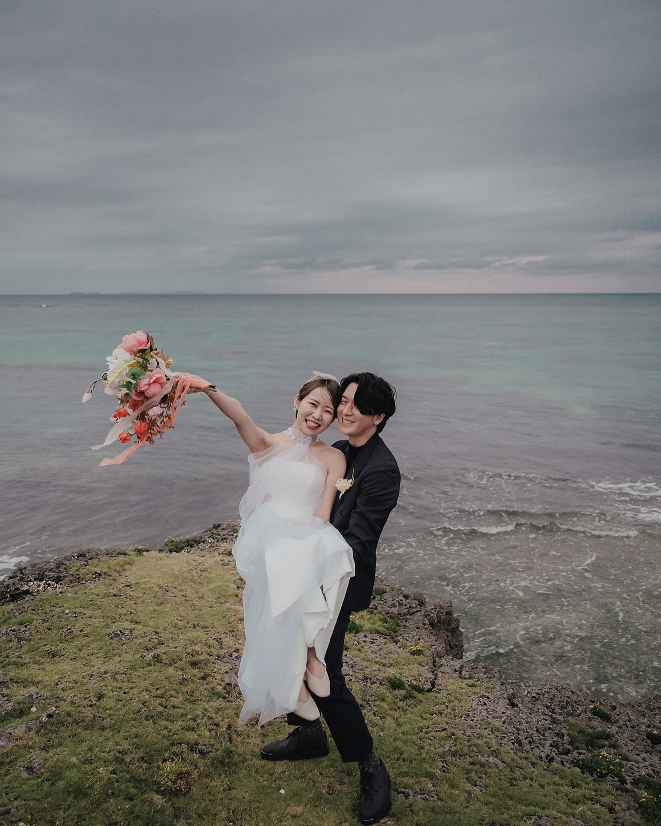 Groom carrying his bride by the emerald sea under cloudy Okinawa skies