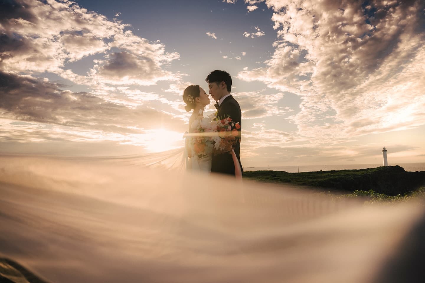 夕焼け空の下、なびくベール越しに向き合う新郎新婦、沖縄の岬にて