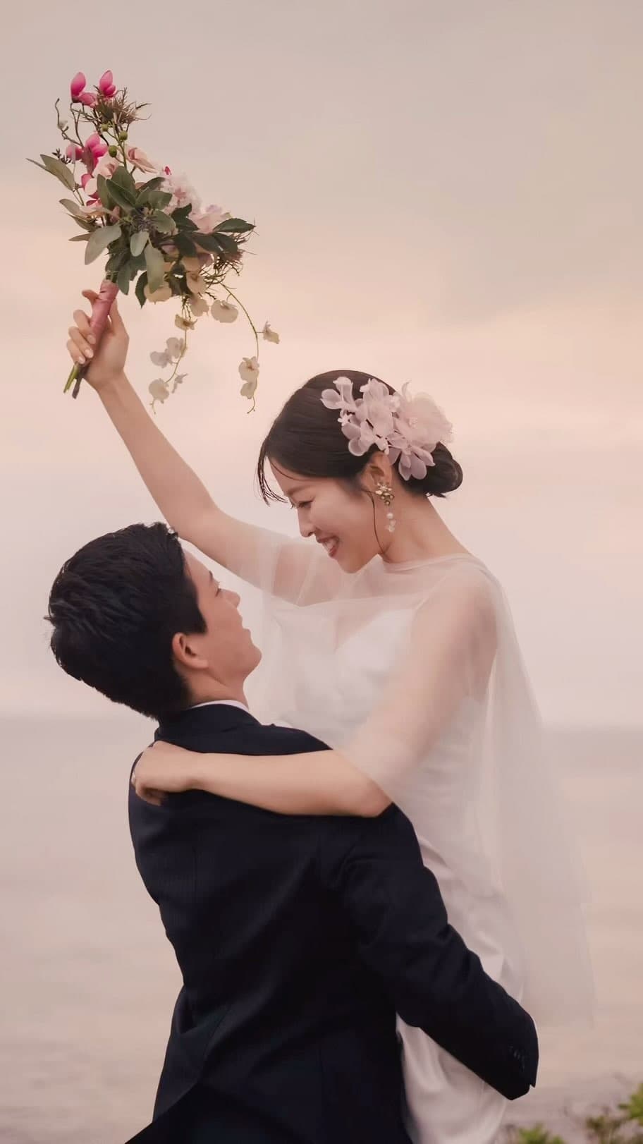 Bride embracing the groom and raising a pink bouquet under a pale rosy sky in Okinawa